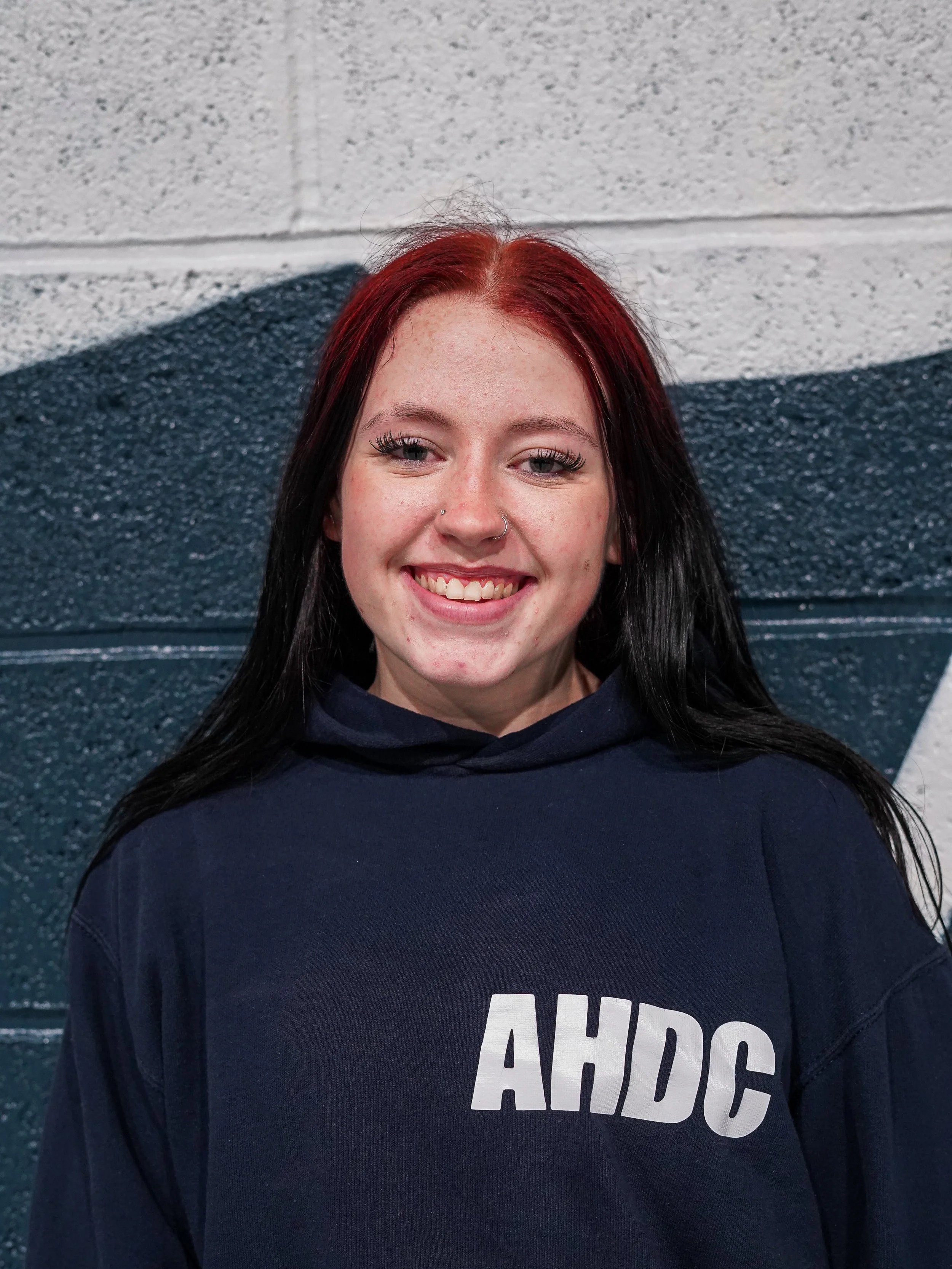 Group of young women in blue T-shirts and sweatpants, sitting cross-legged on the floor, stretching with their hands behind their heads in a dance or yoga class.