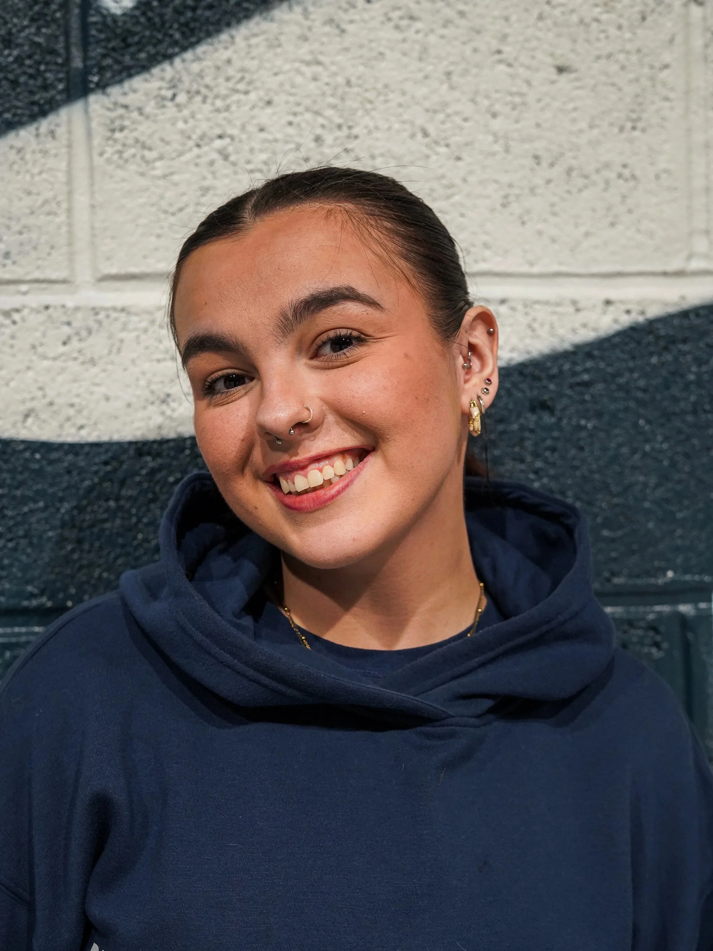 A smiling woman with long brown hair and a ponytail, wearing earrings, is hugging a young girl with red hair in a ponytail. Both are wearing blue shirts. In the background, there are children participating in an activity in a large indoor space with metal walls.