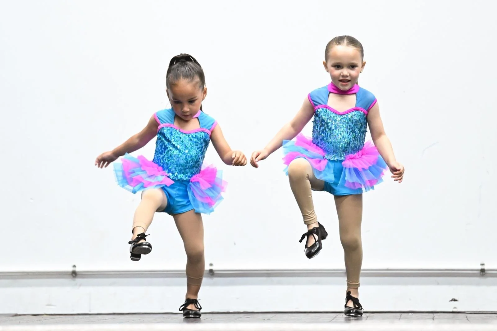 Two young girls in colorful ballet costumes performing a dance on stage, raising one leg.