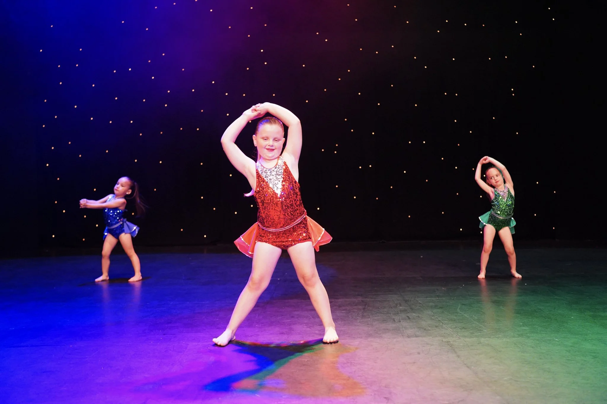 Three young girls are performing a dance routine on stage, wearing colorful sequined costumes, with a dark backdrop decorated with small lights.