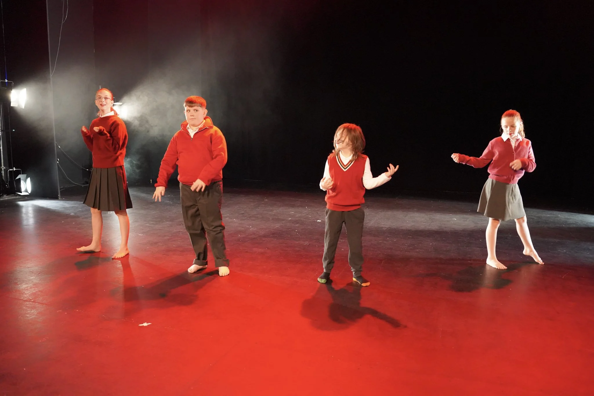 Four children in red tops and skirts or pants standing on stage with black background, some barefoot, under bright stage lights.