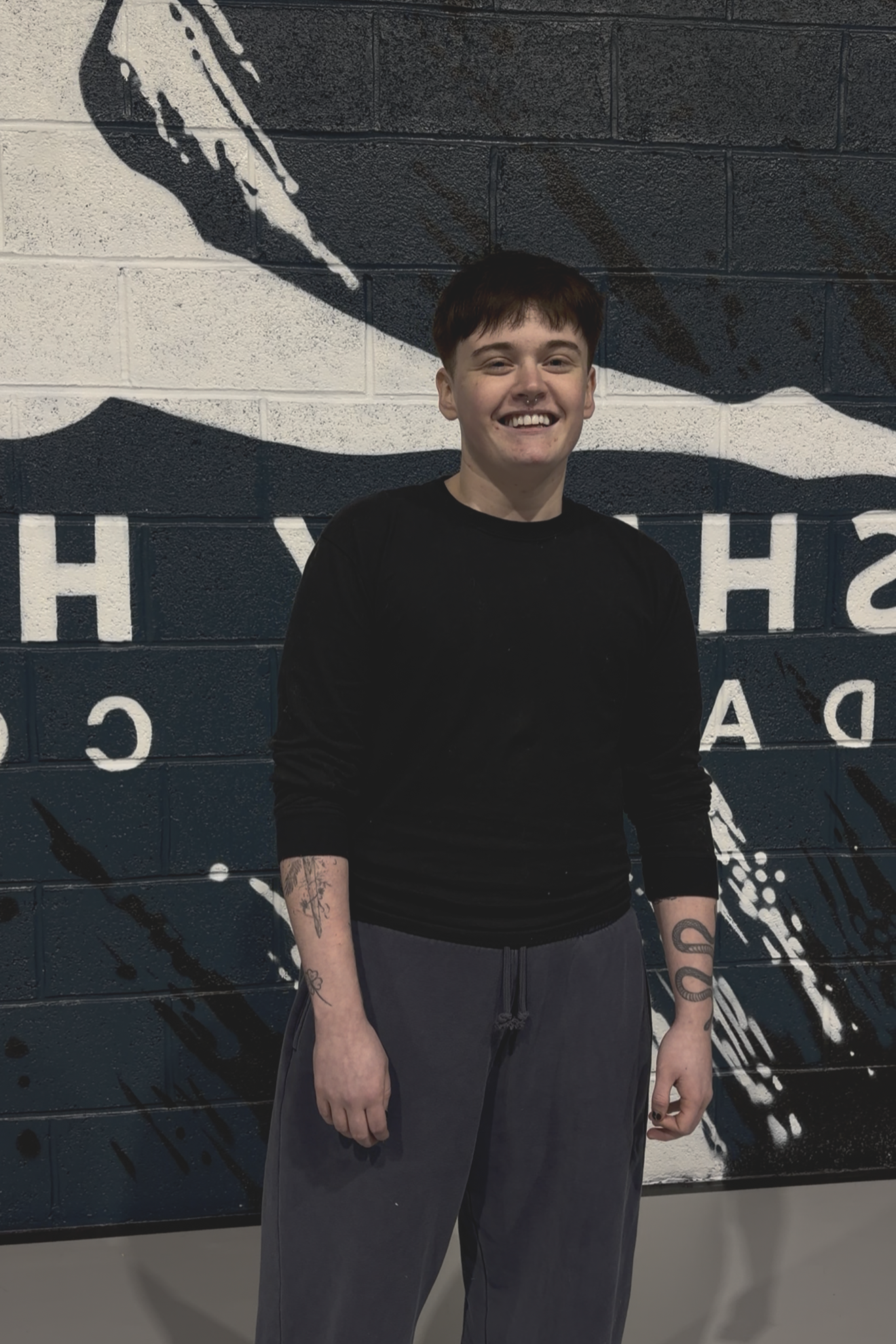 A young girl and a young man stand together in a gym, smiling at the camera. They are wearing matching blue shirts, and the girl has her arm around the man's waist. The gym background includes a black wall with large white lettering.