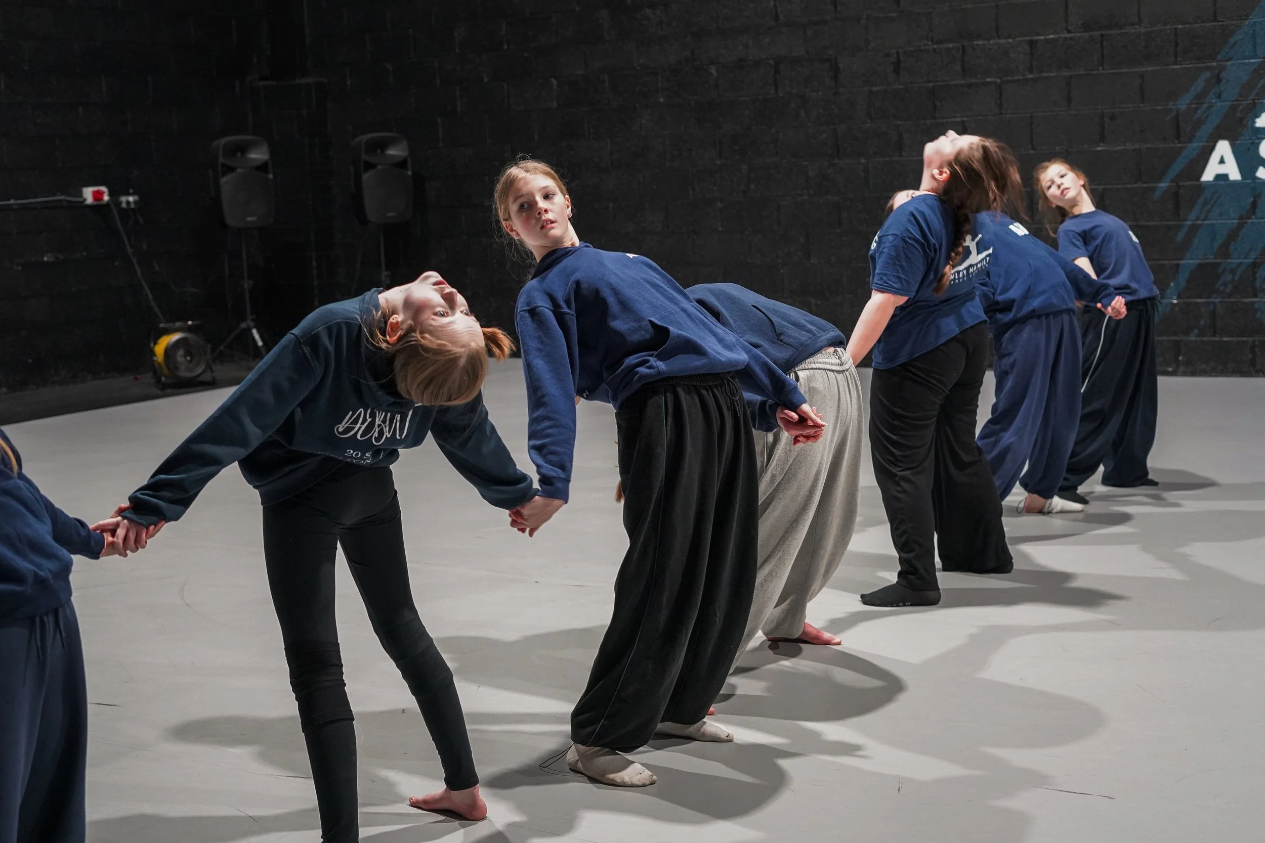 Group of young girls holding hands and leaning back in a dance or theater rehearsal on a gray studio floor with black walls.