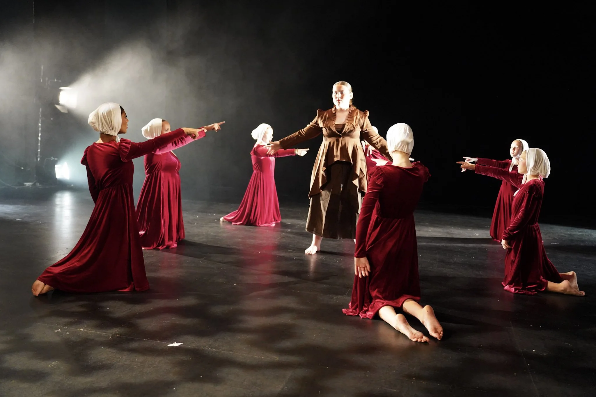 Ballet performers on stage, with one woman in the center wearing a brown costume and six girls dressed in red dresses with white head coverings, striking dance poses.