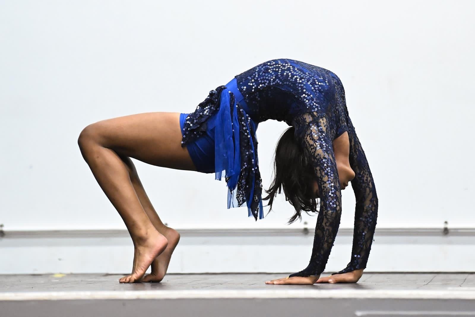 A woman in a blue sequined and lace costume performing a yoga pose on a stage with a plain white background.