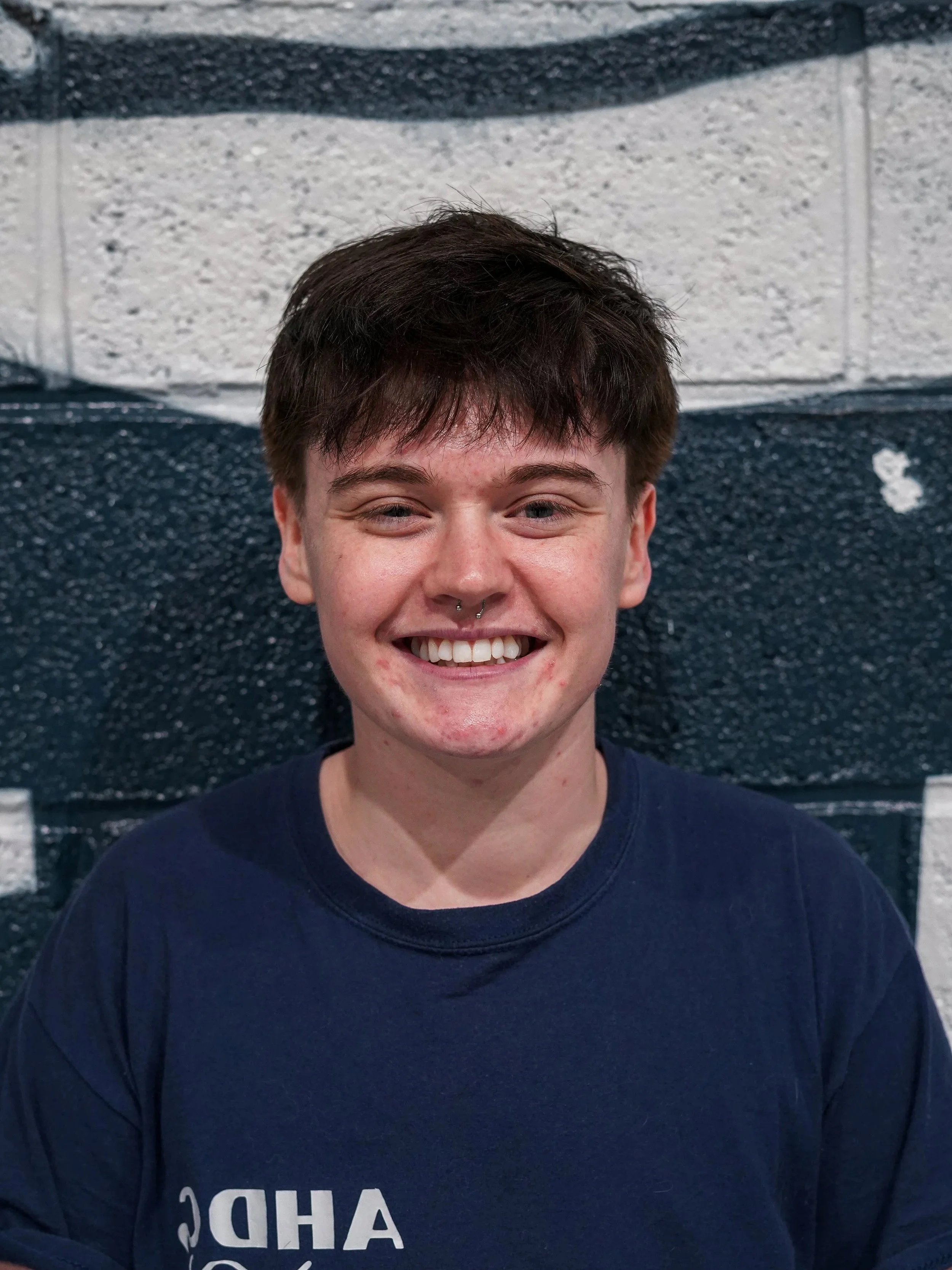A young girl and a young man stand together in a gym, smiling at the camera. They are wearing matching blue shirts, and the girl has her arm around the man's waist. The gym background includes a black wall with large white lettering.