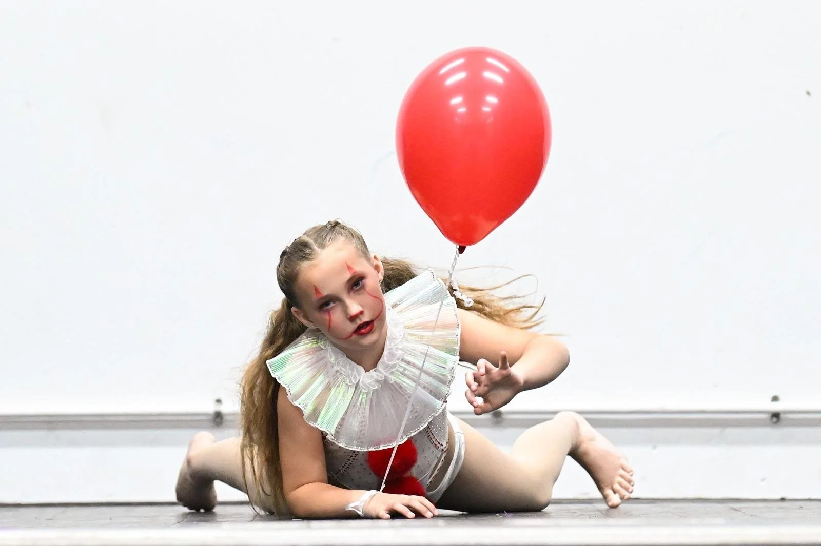 A young girl dressed as a clown or mime, sitting on the floor with a red balloon, wearing theatrical makeup, a ruffled collar, and beige tights, against a plain white background.