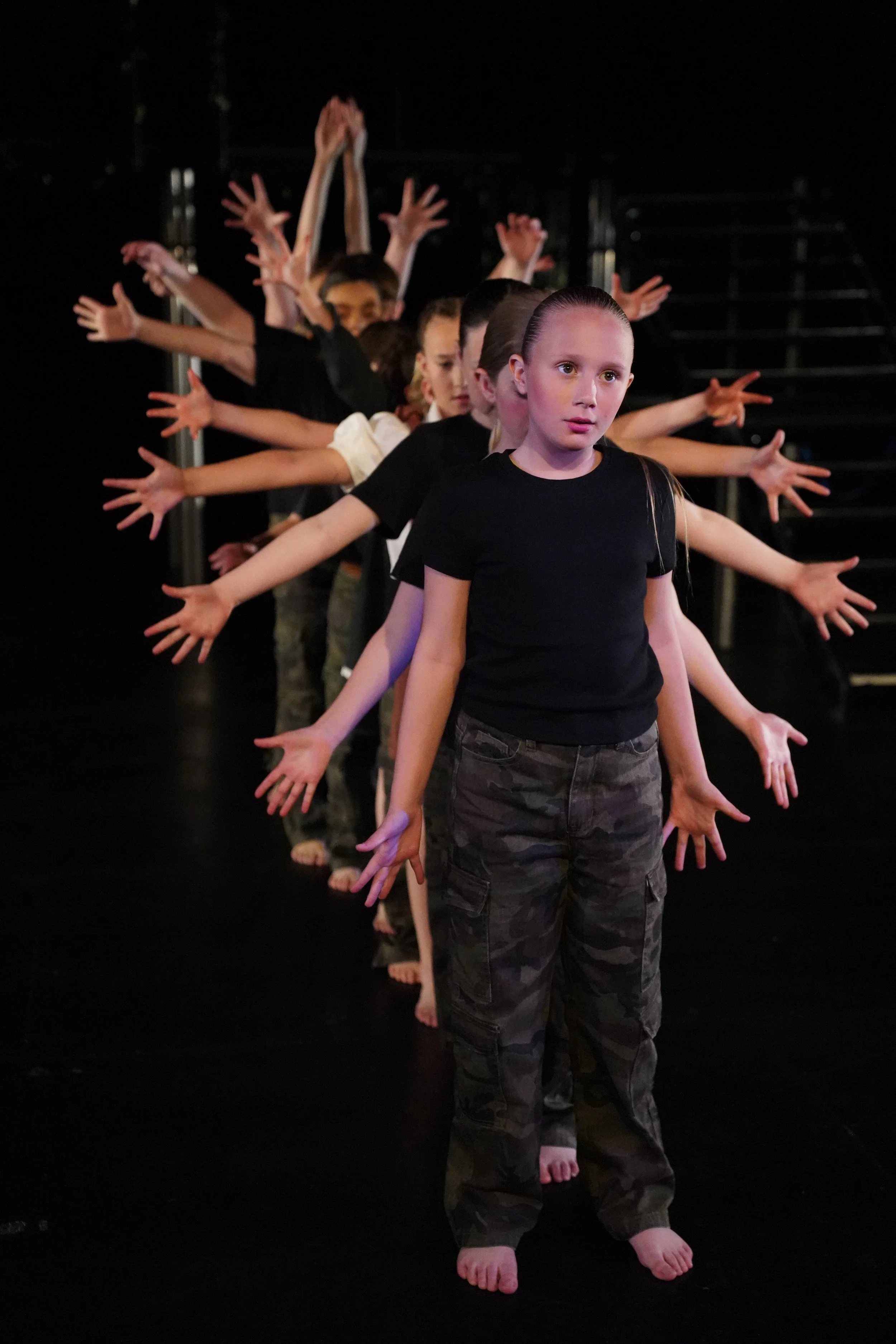 Young children standing in a line with their arms extended and fingers spread, practicing a dance or theater performance on a dark stage.