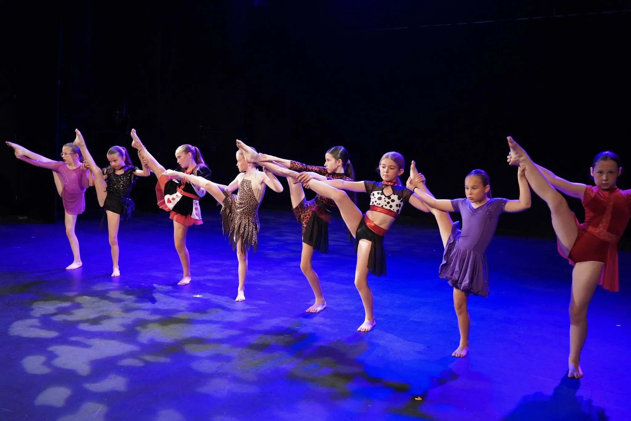 Young girls dressed in colorful costumes performing a ballet dance on stage, with some holding poses and others stretching in a dance routine, under dark stage lighting.