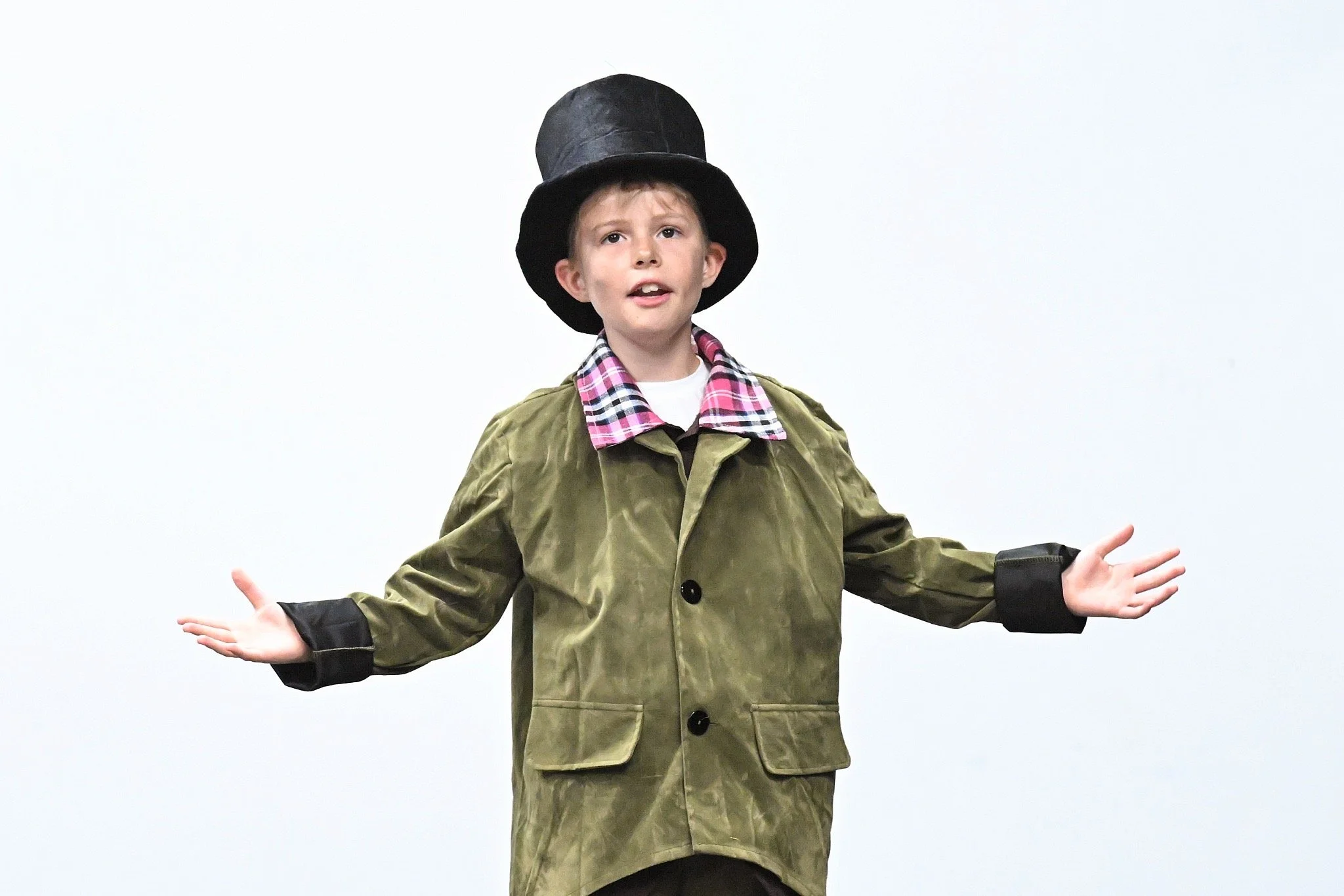 A young boy is standing against a plain white background, wearing a large black top hat, an olive-green coat with black cuffs, and a pink plaid shirt. He has his arms outstretched with his palms facing up.