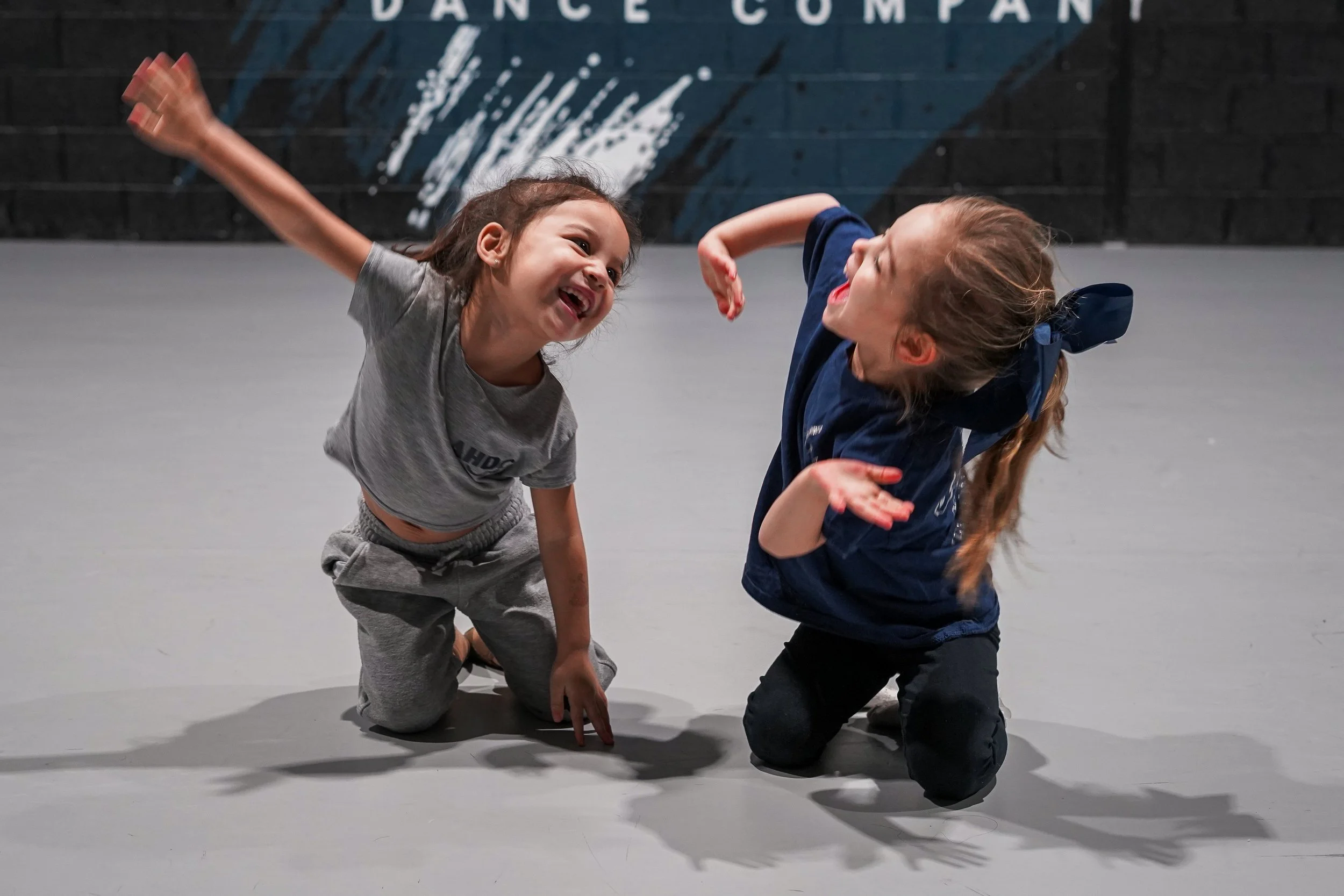 Two young girls are playing and laughing on the floor of a dance studio, with a blue and black wall in the background.