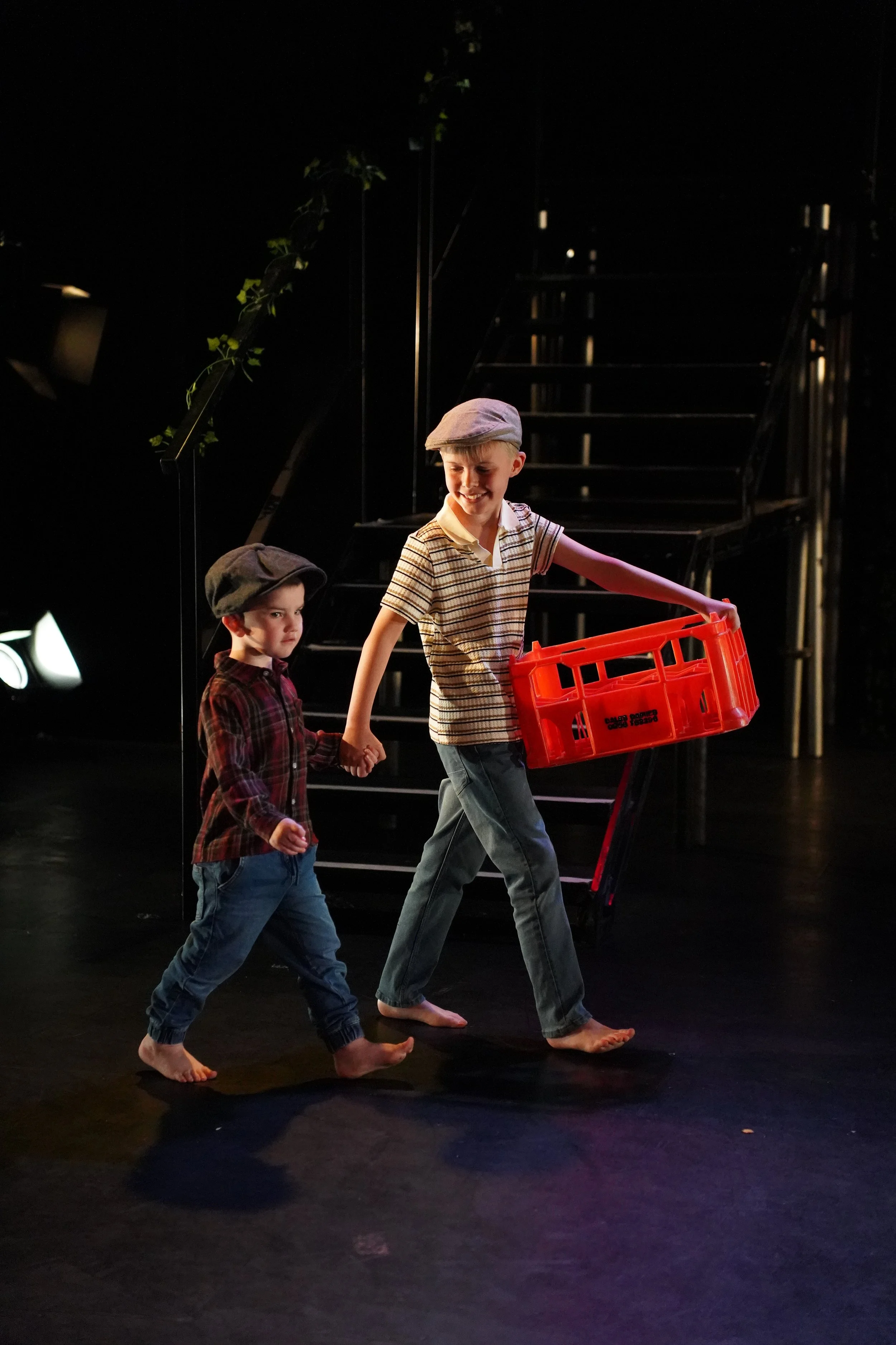 Two young boys walking barefoot on a stage, holding hands, with one boy carrying an orange crate. They are wearing vintage-style newsboy caps and casual clothing. The background is dark with a staircase and some greenery.