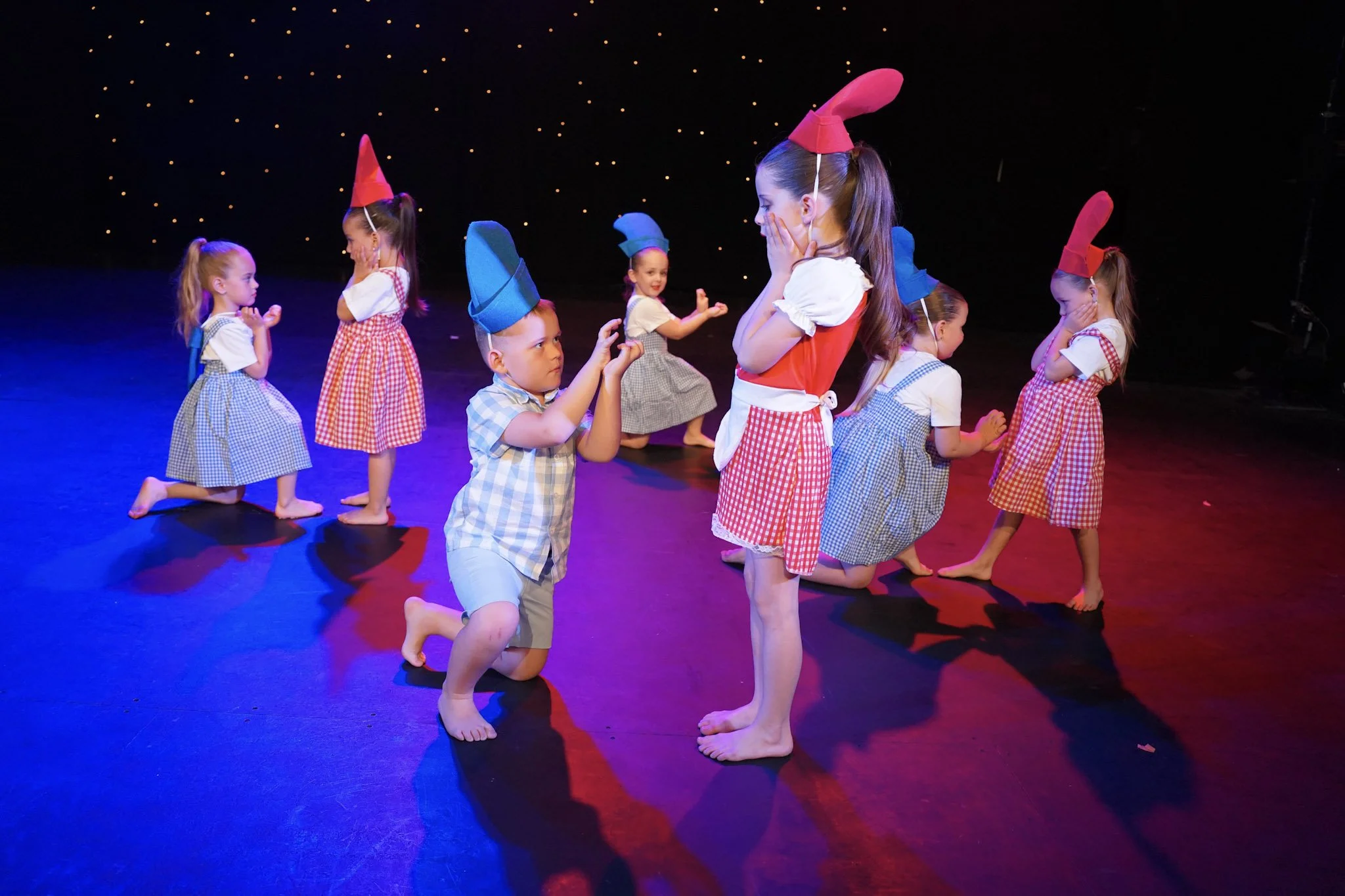 Children dressed as elves with red and blue hats perform a dance on stage under colorful lighting.
