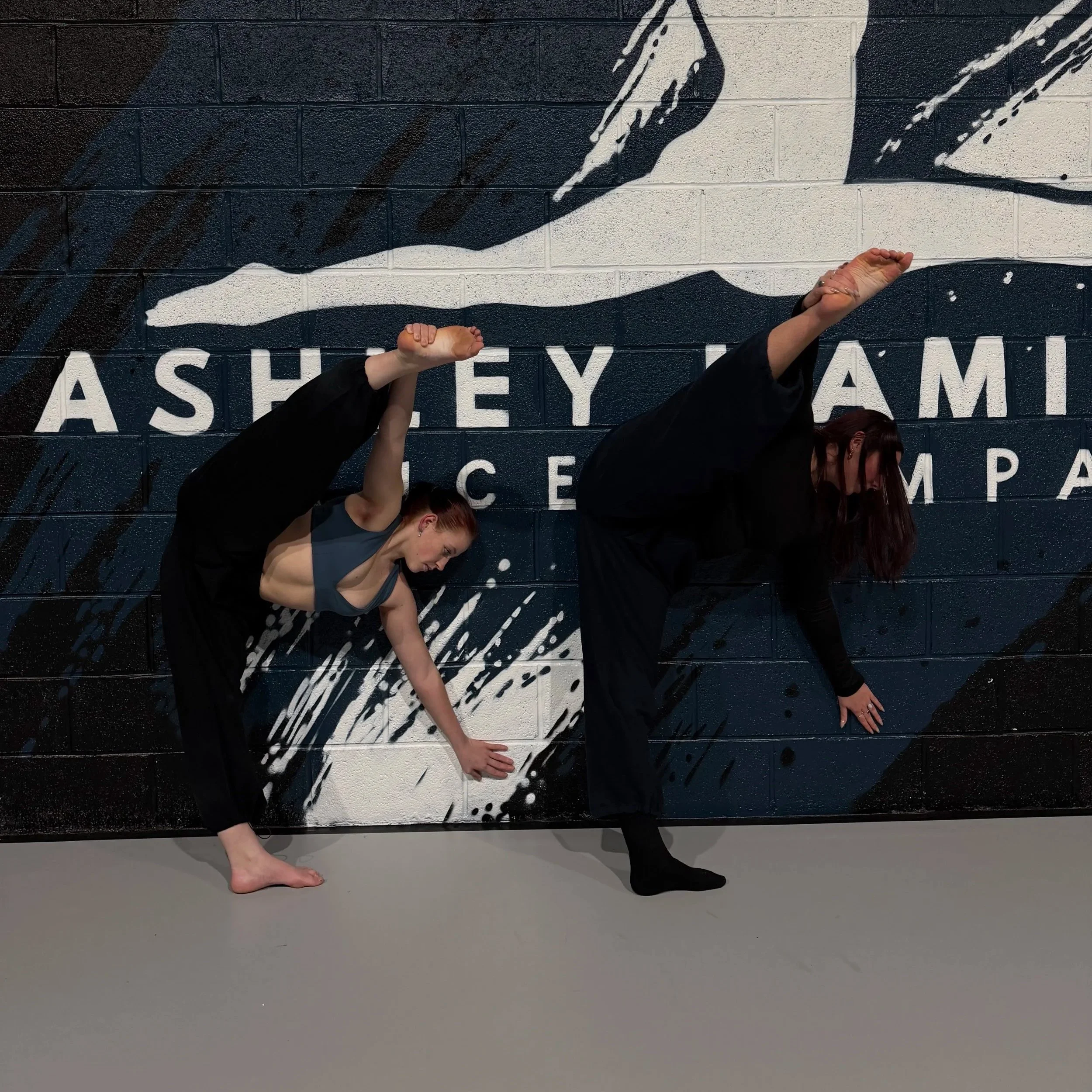 Group of young women in blue T-shirts and sweatpants, sitting cross-legged on the floor, stretching with their hands behind their heads in a dance or yoga class.