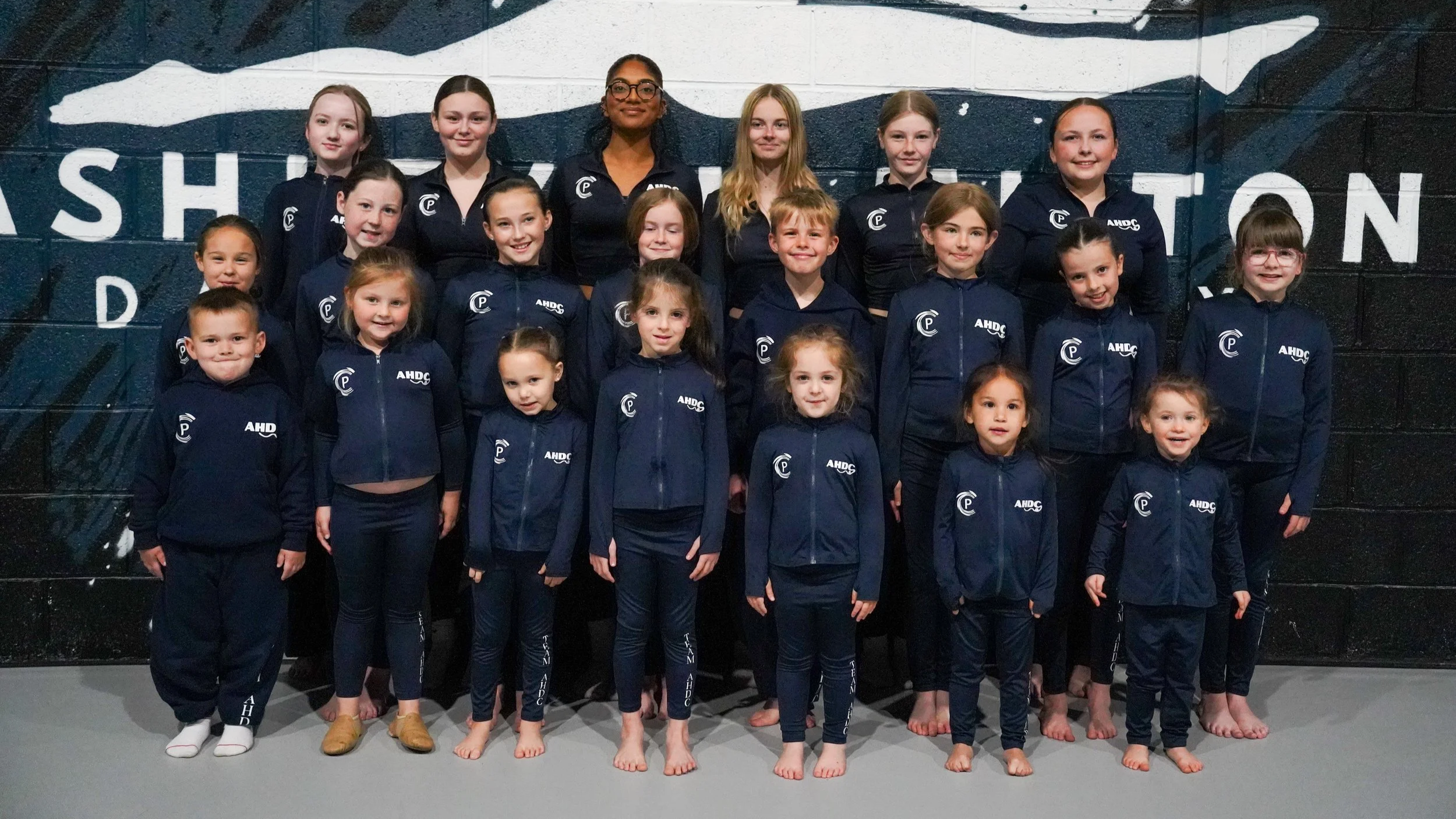 Group photo of young girls and boys in matching navy blue athletic uniforms with the letters 'AHDC' and a logo, standing on a gymnasium floor in front of a wall with mural art and an abstract black-and-white design.