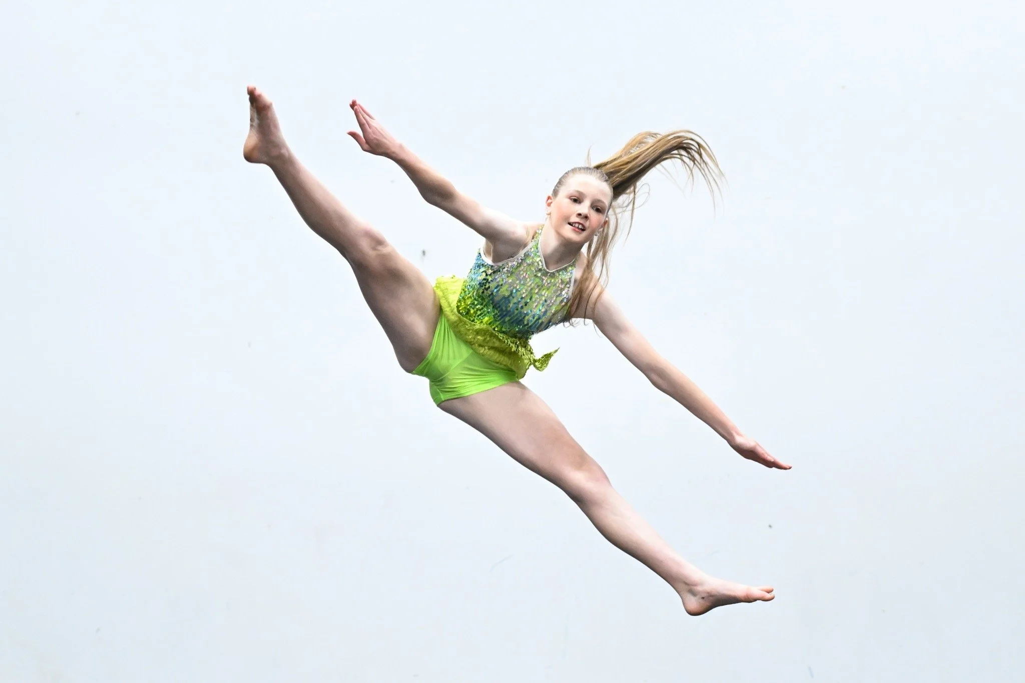 A young girl performing a gymnastics pose mid-air against a plain, light-colored background.