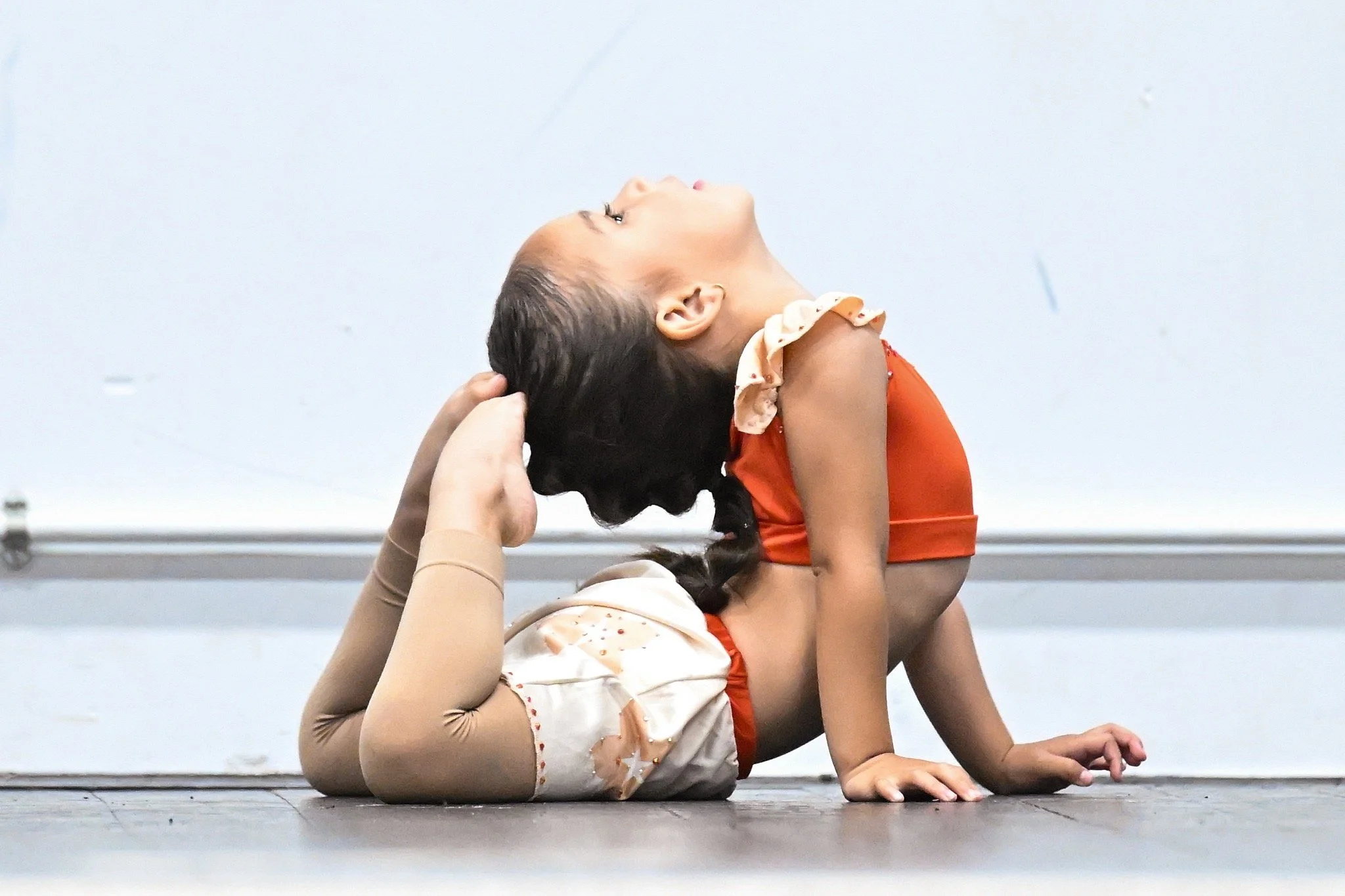 Young female gymnast performing a backbend on the floor with an instructor helping her, against a plain white wall.