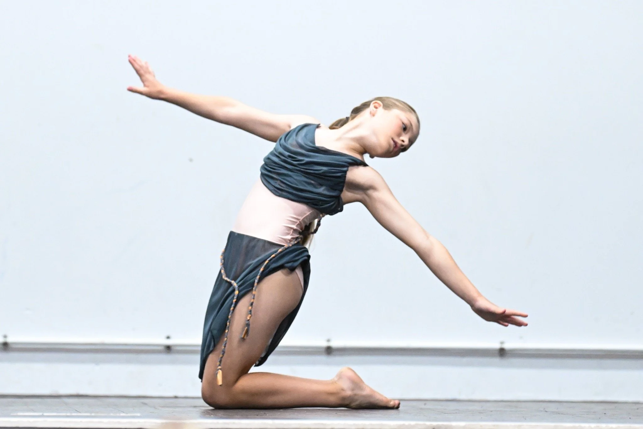 A young female dancer performing a contorted pose on stage, with arms extended and body bent forward, against a plain light background.