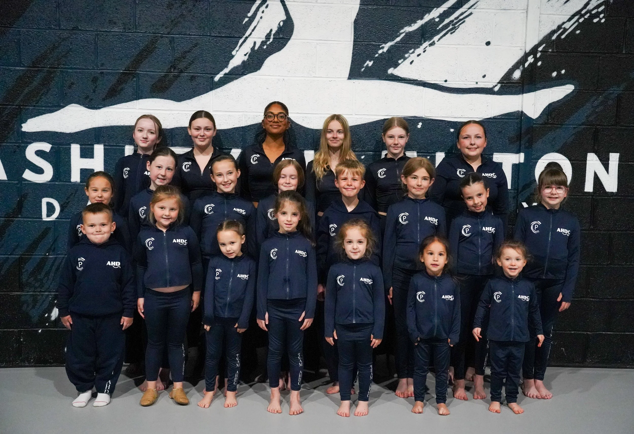 A group of children and a young woman, likely a coach, standing in front of a wall with a large mural of a hand pointing, on a black and white background. The children are dressed in matching navy blue jackets and pants with the logo 'AHDC'.