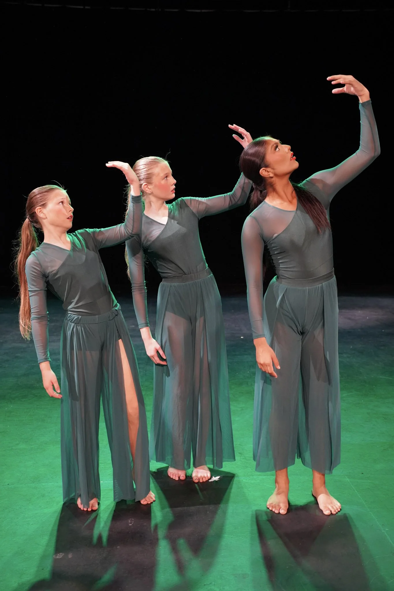 Three female ballet dancers on stage, wearing matching sheer gray dance costumes, performing a synchronized dance with their arms raised and gazing upward, with one dancer using a balance board.