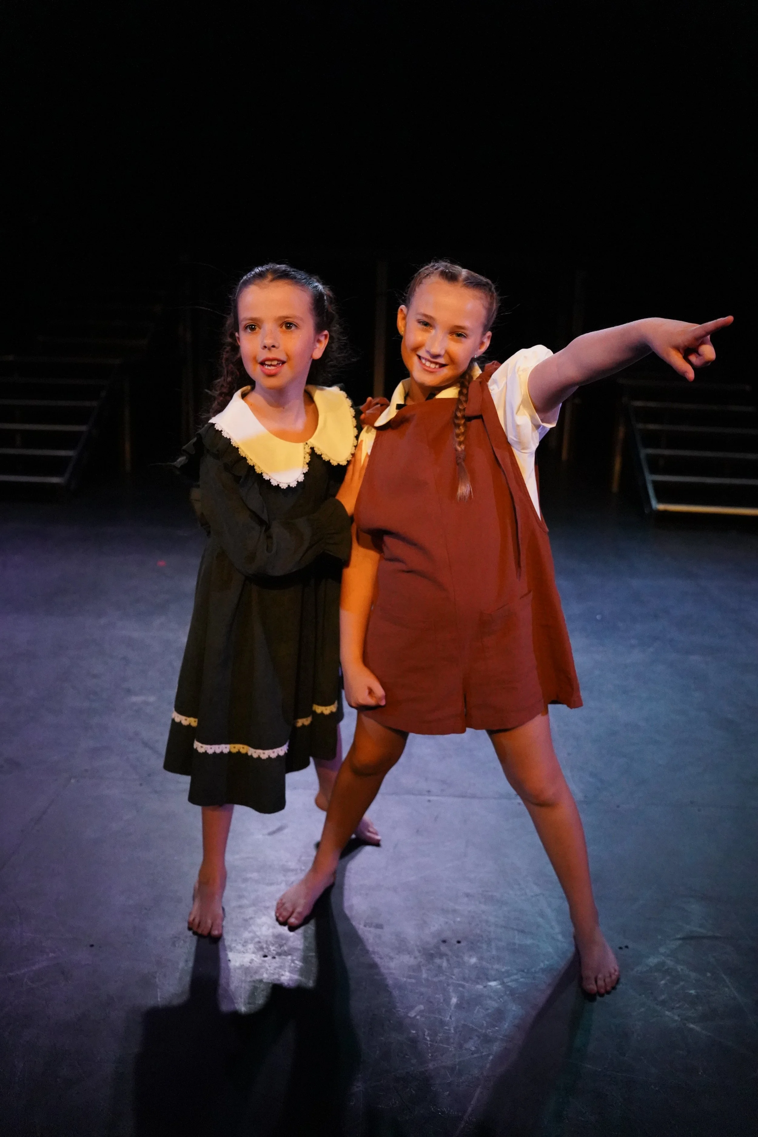Two young girls with braided hair, one in a black dress with white lace collar and trim, and the other in a brown dress with a beige shirt, stand barefoot on a dark stage with black steps behind them. One girl is pointing outward with a smile, the ot