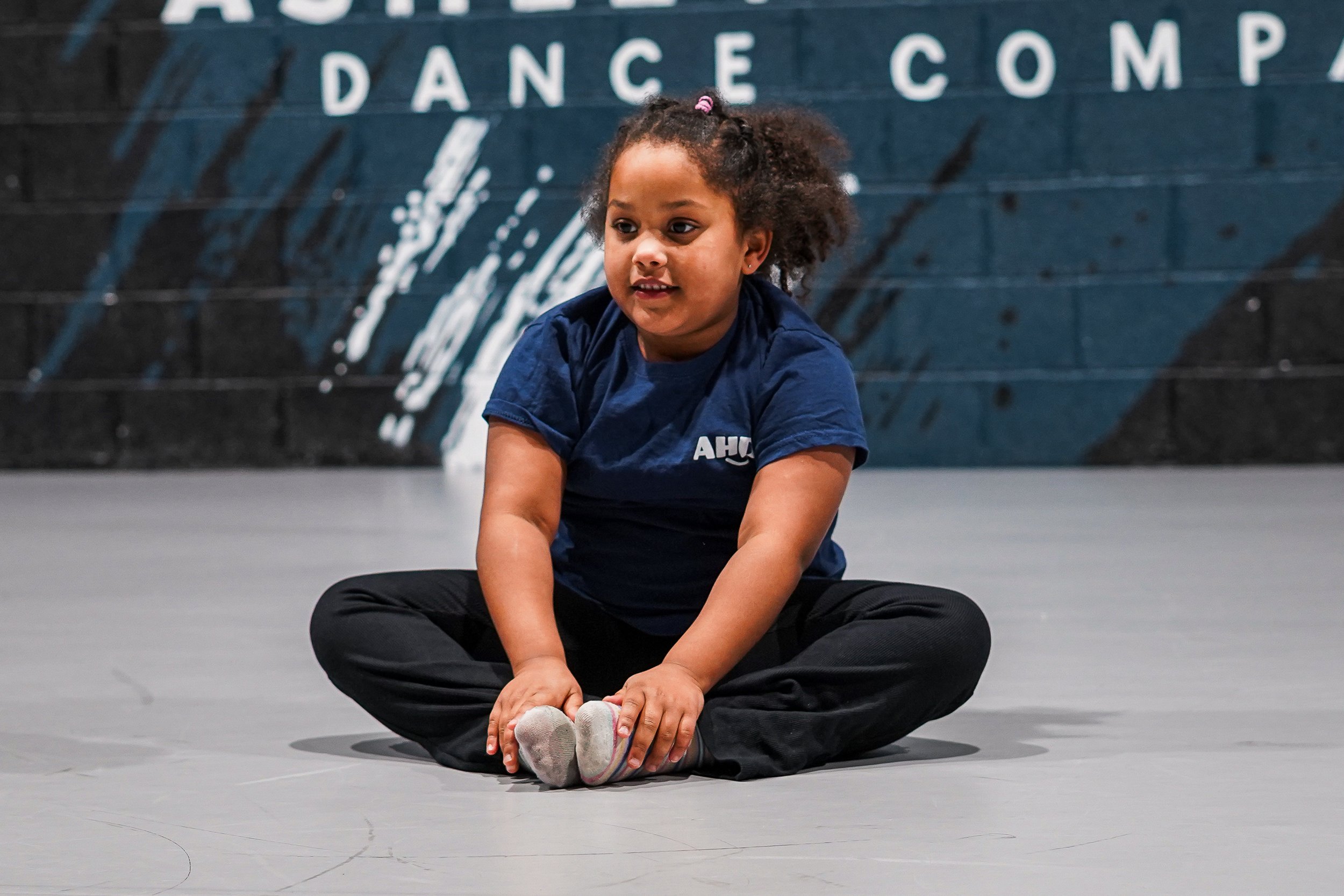A young girl sitting cross-legged on a dance studio floor, holding her toes, with a large dance studio mural in the background.
