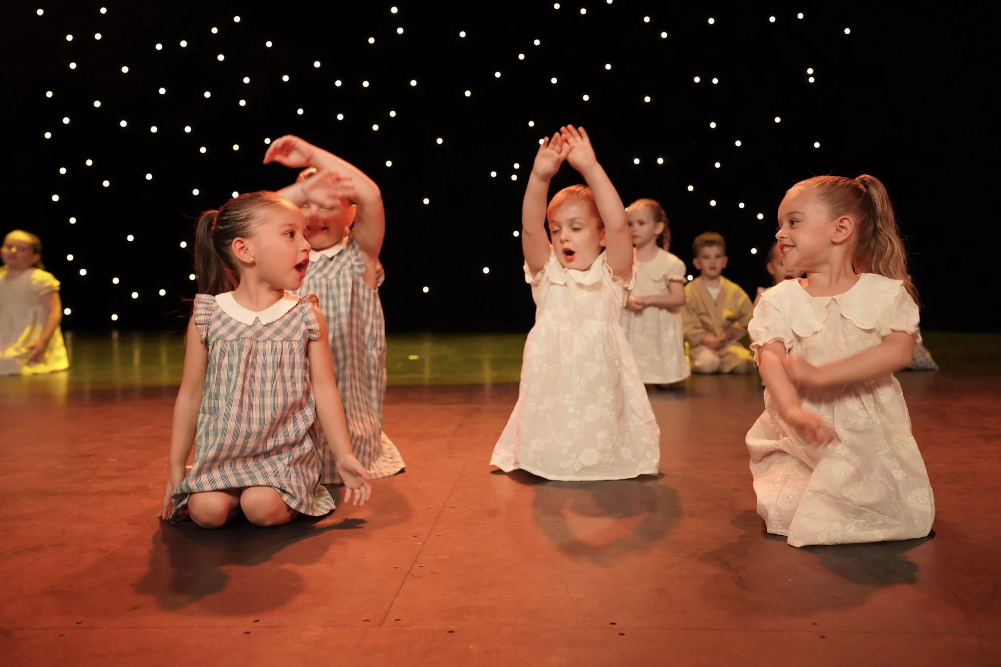 Group of young children dressed in vintage-style clothing performing a dance or song on a stage with starry background lighting.