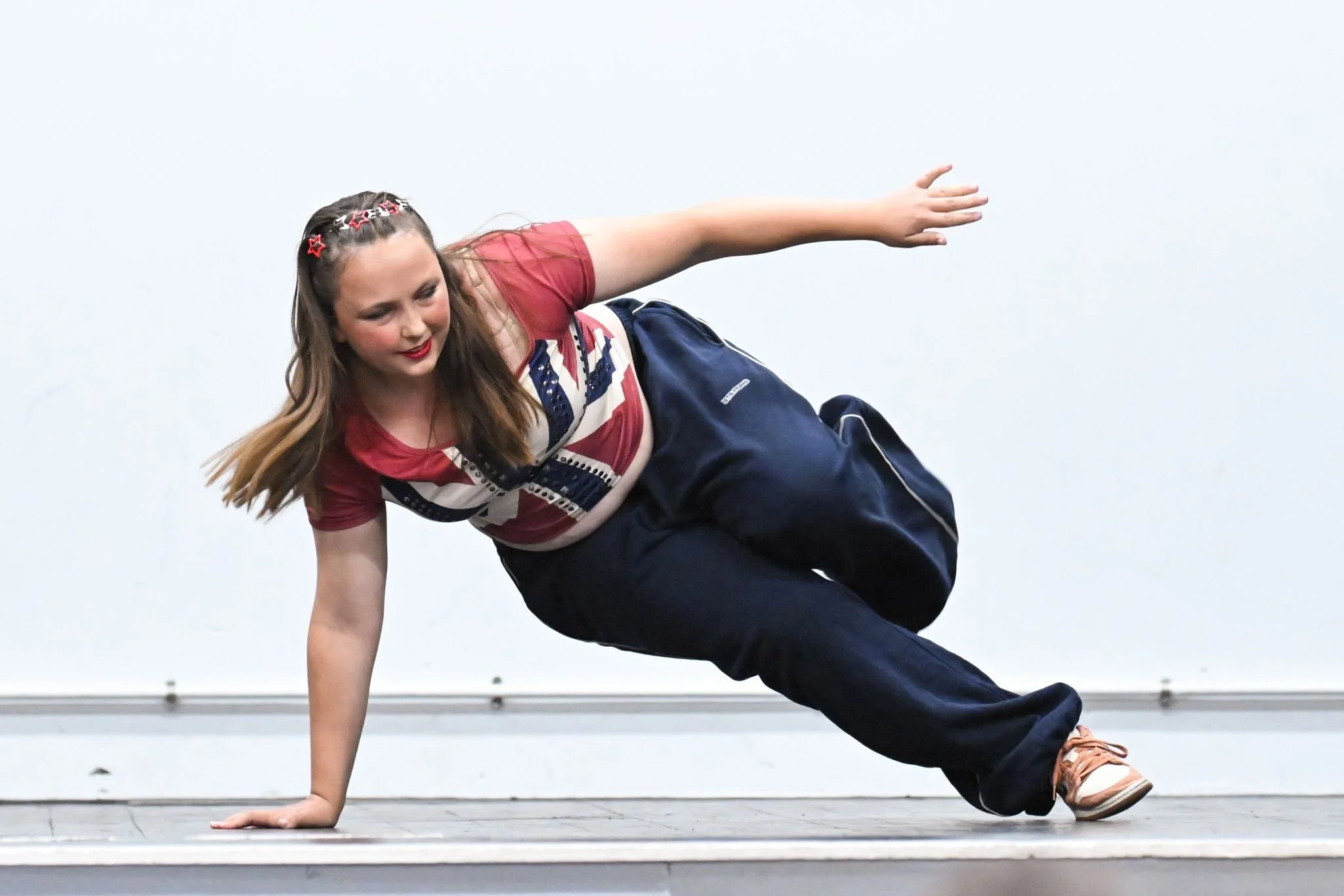Young woman in casual clothing breakdancing on stage, balancing on one hand with her legs extended and her hair loose, against a plain background.