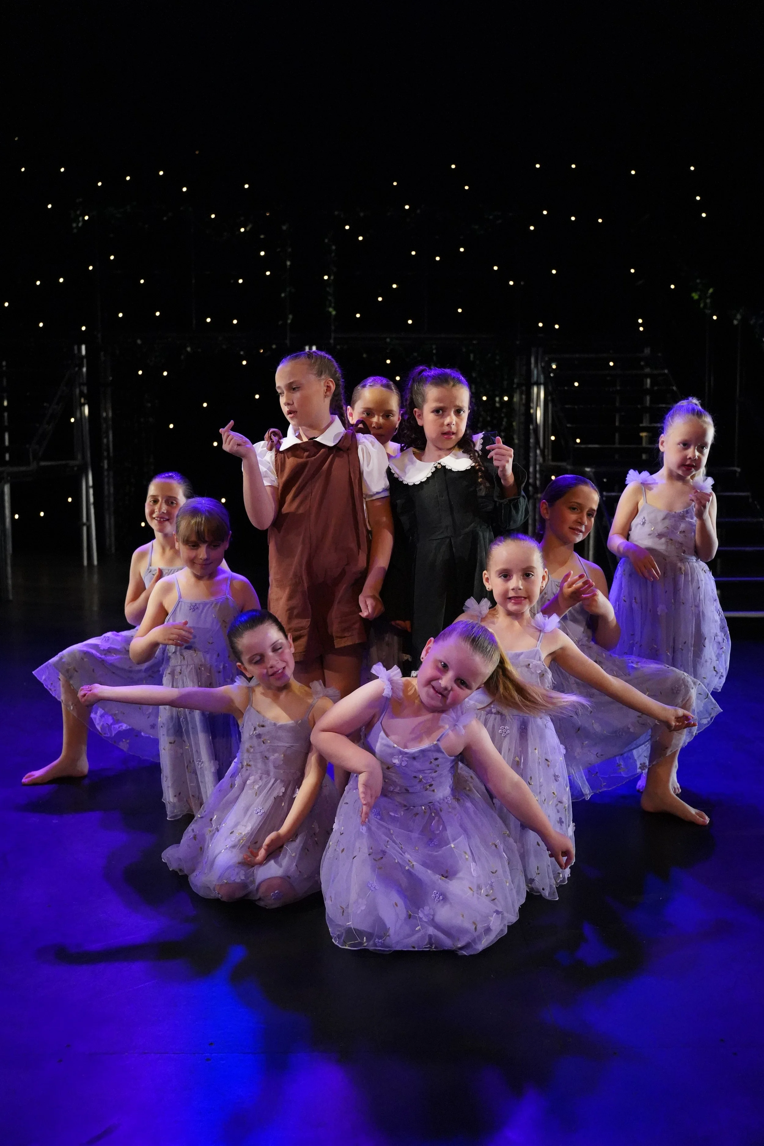 Children in ballet costumes performing on stage with dark background and small lights, some kids standing, others sitting or lying on the floor, striking various poses.
