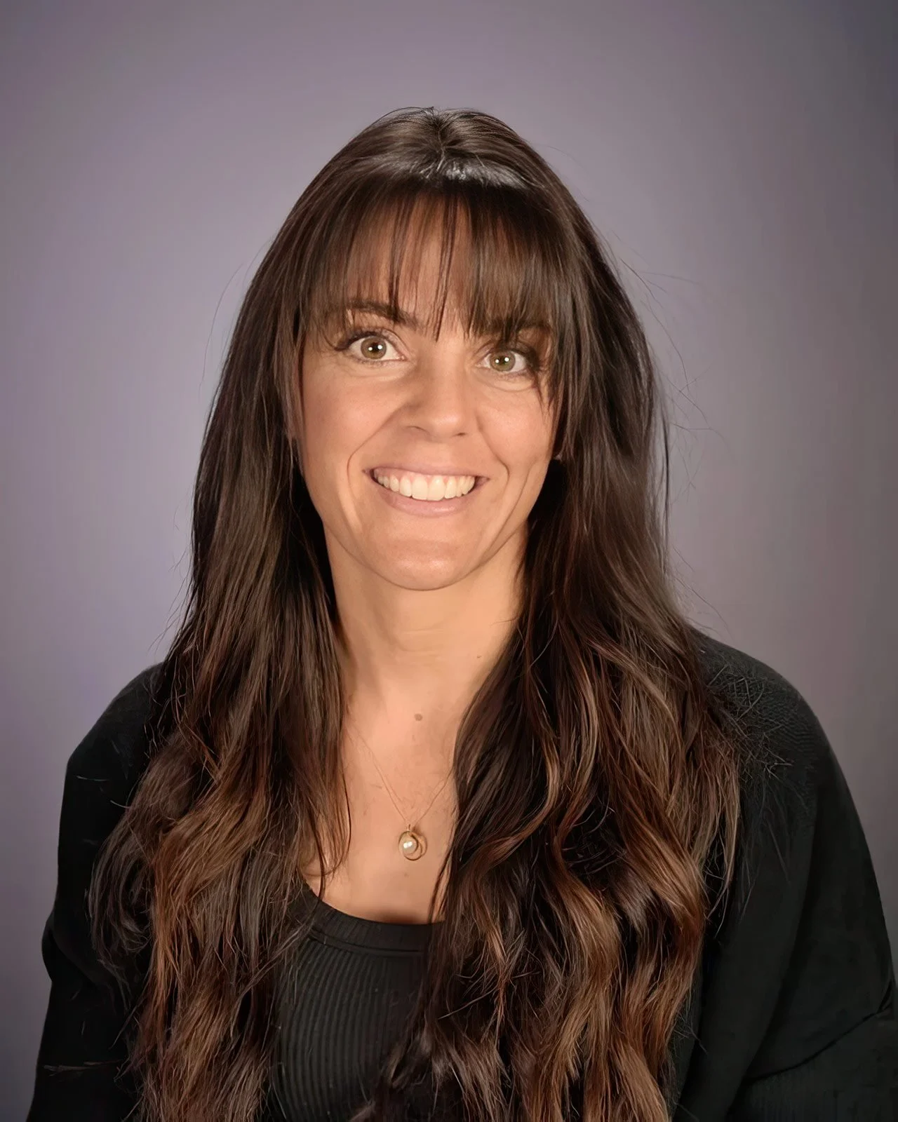 Portrait of a woman with long brown hair, wearing a black top and a necklace with a pearl pendant, smiling against a gray background.