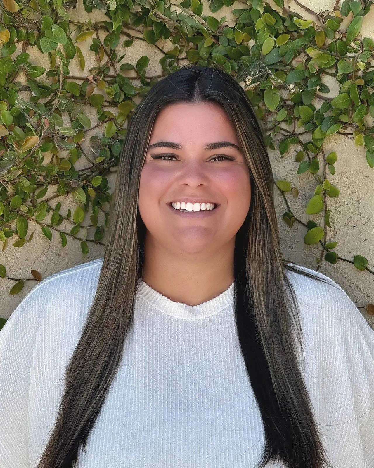 Young woman with long brunette hair smiling in front of a green leafy background.