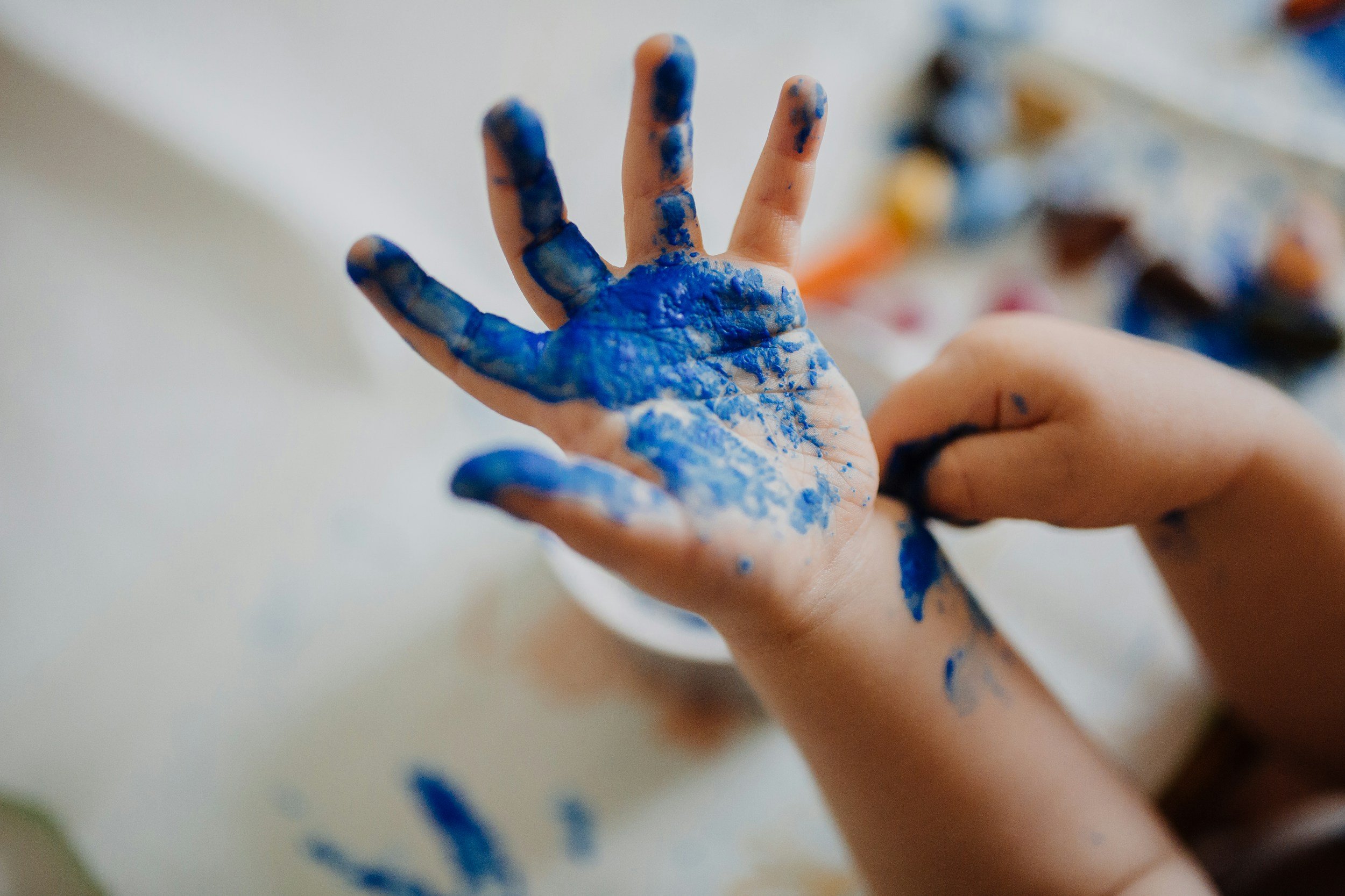 A child's hands covered in blue and black paint, with some paint on the table surface.