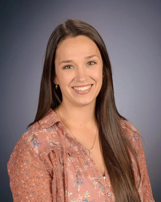 A young woman with long brown hair, smiling, wearing a floral blouse and a gold necklace, posed against a solid dark background.
