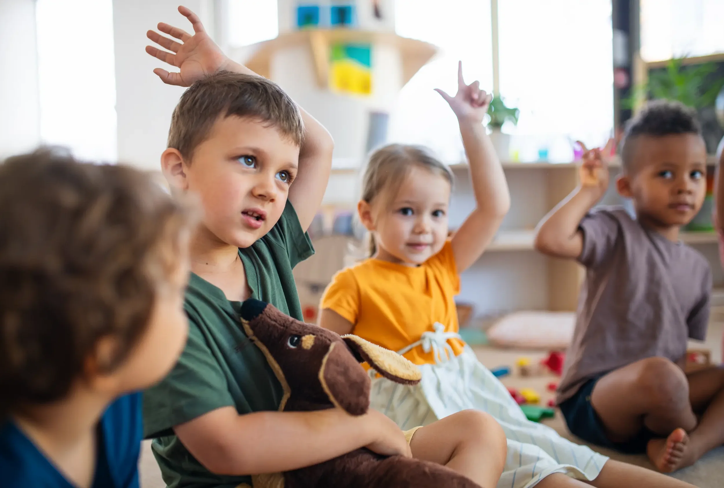 Four young children sitting on the floor in a classroom, raising their hands to answer a question.