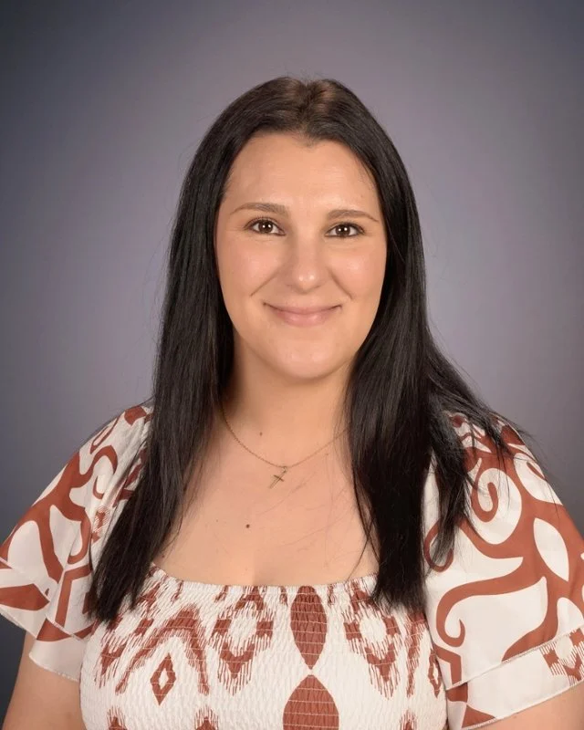 A woman with long dark hair and fair skin, smiling at the camera, wearing a white and brown patterned top and a delicate gold cross necklace against a neutral background.