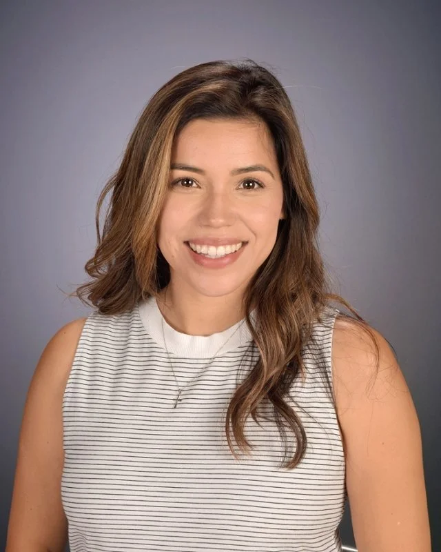 A smiling woman with shoulder-length brown hair wearing a white sleeveless top with black stripes, posing against a gray background.