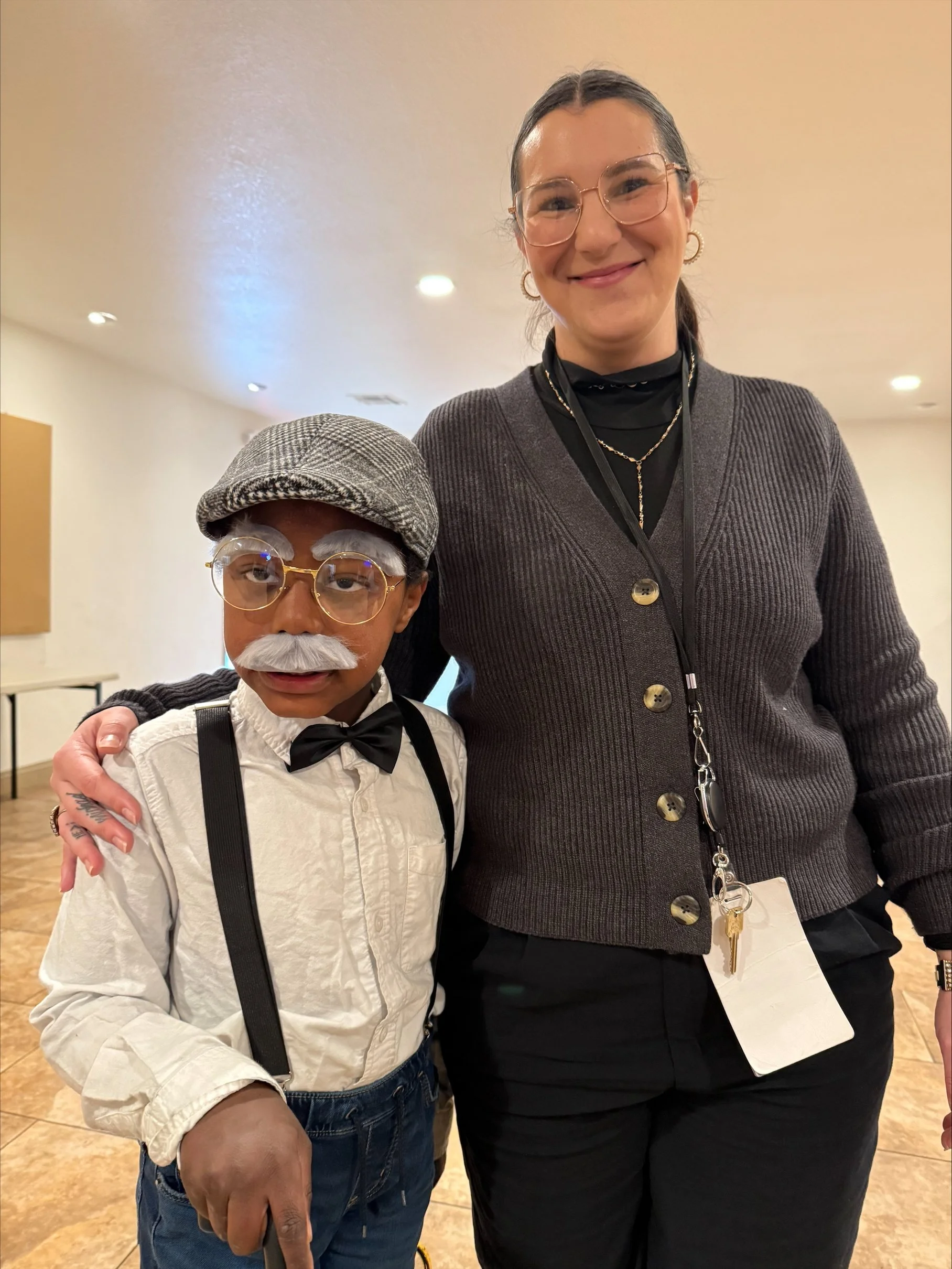 A young boy dressed in vintage style, wearing glasses, a gray hat, a white shirt with a black bow tie, suspenders, and a fake white mustache. He is standing next to a woman who is smiling, wearing glasses, a dark cardigan, and has a lanyard with keys