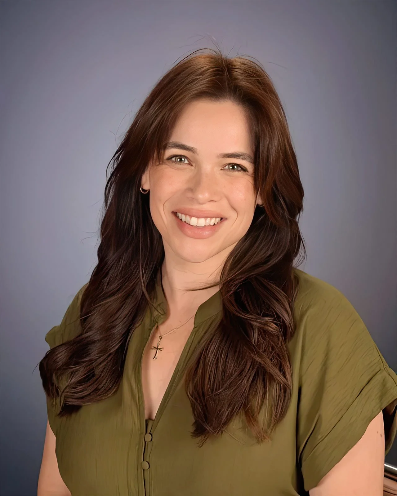 A woman with long, wavy brown hair, smiling, wearing a green blouse and a necklace with an ankh pendant, against a gray background.