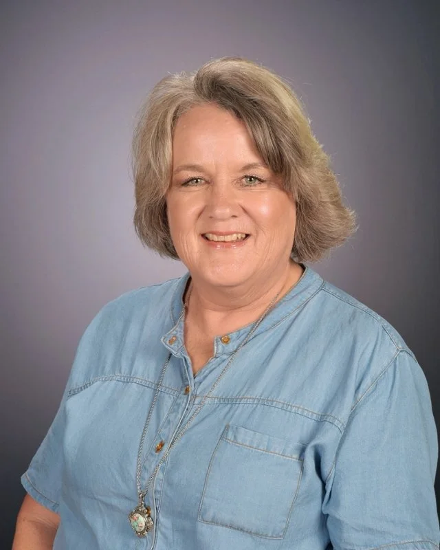 Portrait of a smiling woman with short, gray hair wearing a denim shirt and a necklace, against a gray background.