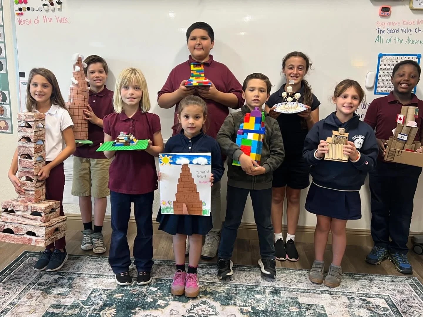 Group of children standing in front of a whiteboard, holding various craft projects, including a brick tower, a rainbow-colored structure, and a drawing of a tower with the label 'Tower of Babel'.