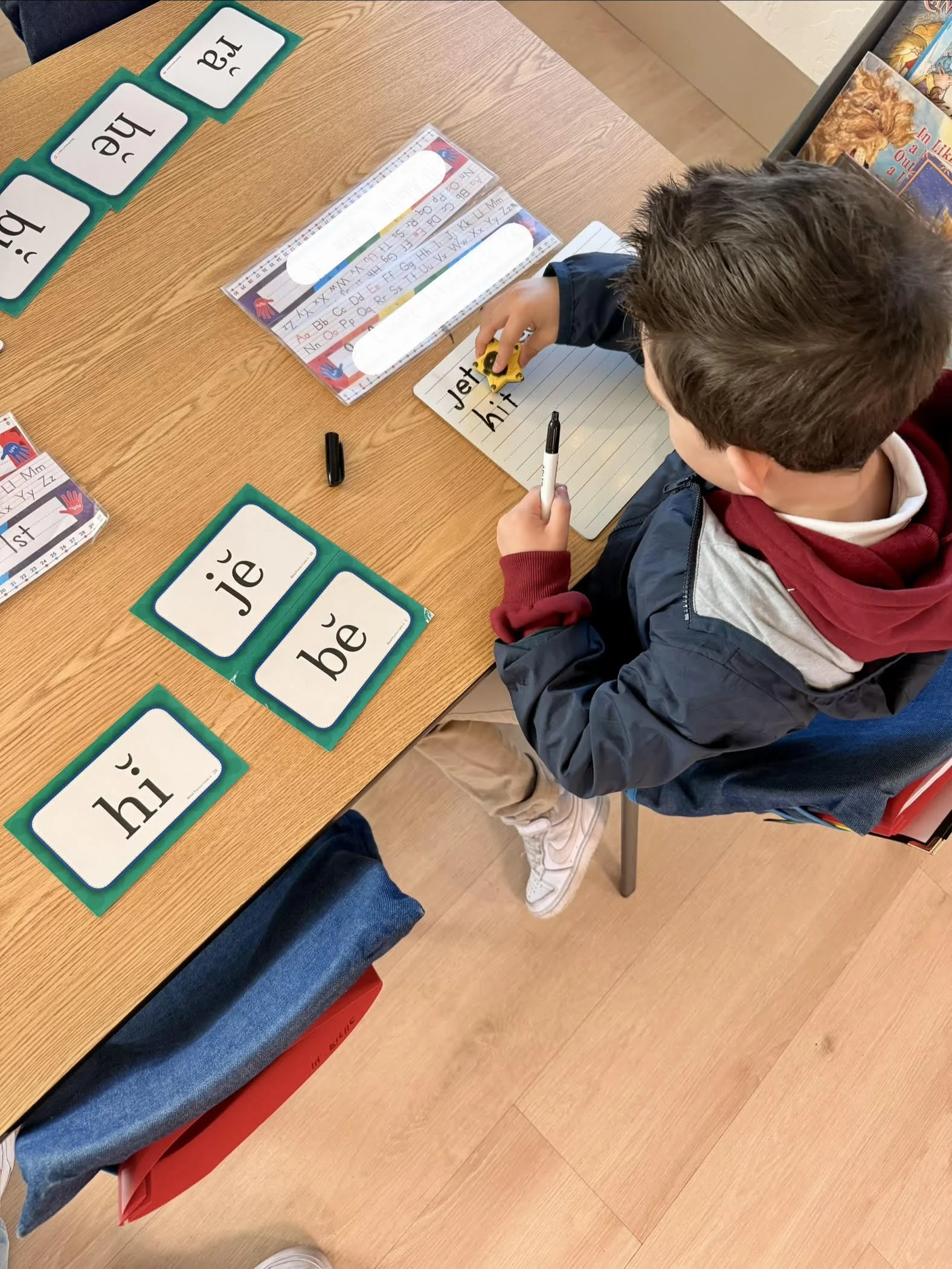 A young boy sitting at a desk practicing writing words with a toy car, surrounded by flashcards with common words like 'hi,' 'je,' 'bée,' and 'la' in a classroom.