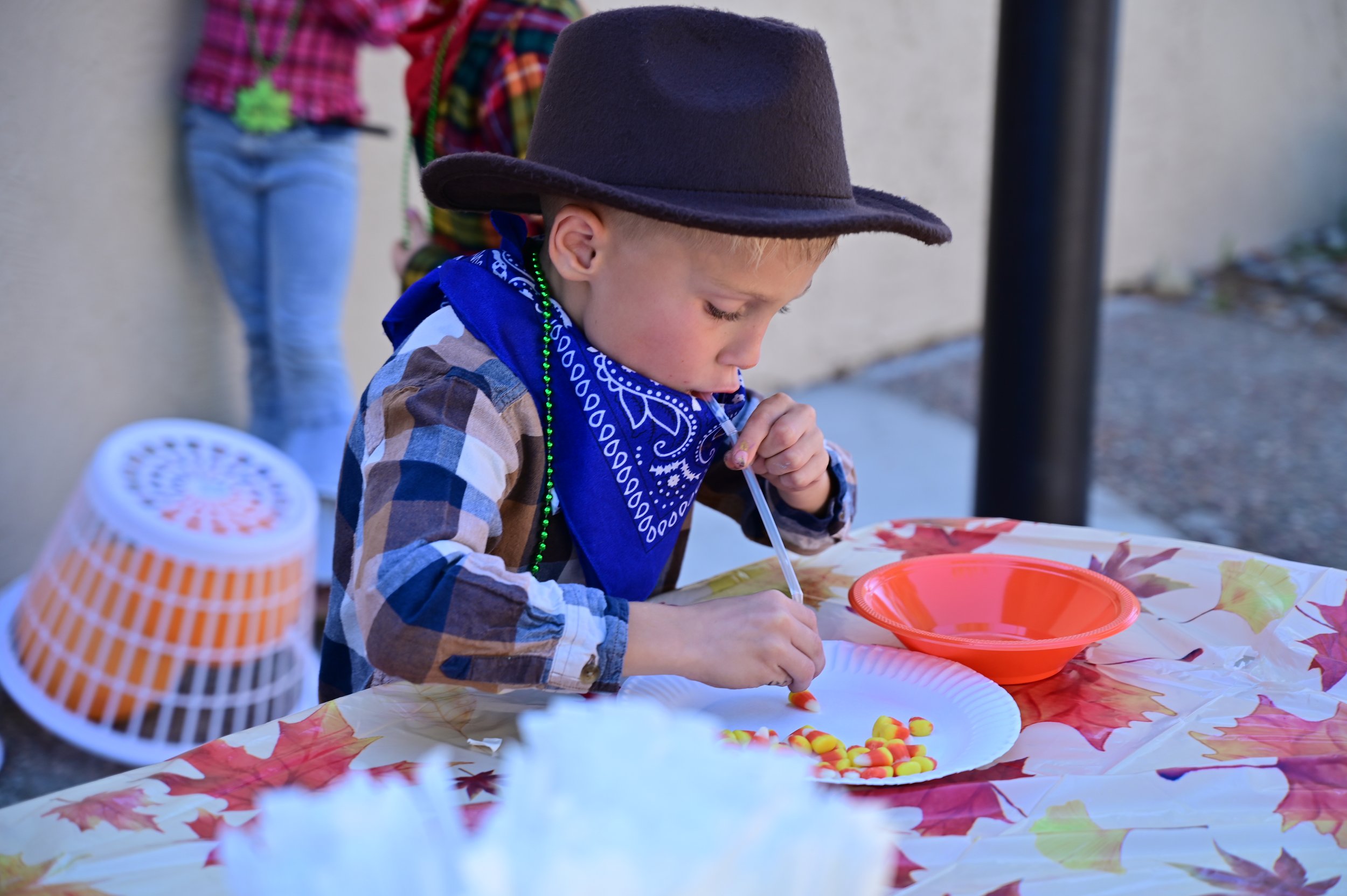 Young boy with a cowboy hat, plaid shirt, and blue bandana, sitting at a table with a colorful autumn-themed tablecloth, eating candy corn from a white paper plate with a blue bowl and orange bowl nearby, outdoors.