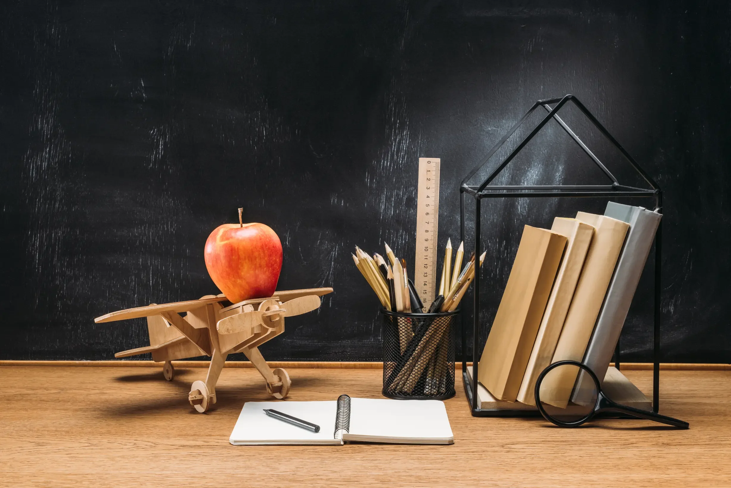 Blackboard with school supplies, including a wooden airplane with an apple on top, a ruler, notebooks, a pen, and a magnifying glass.
