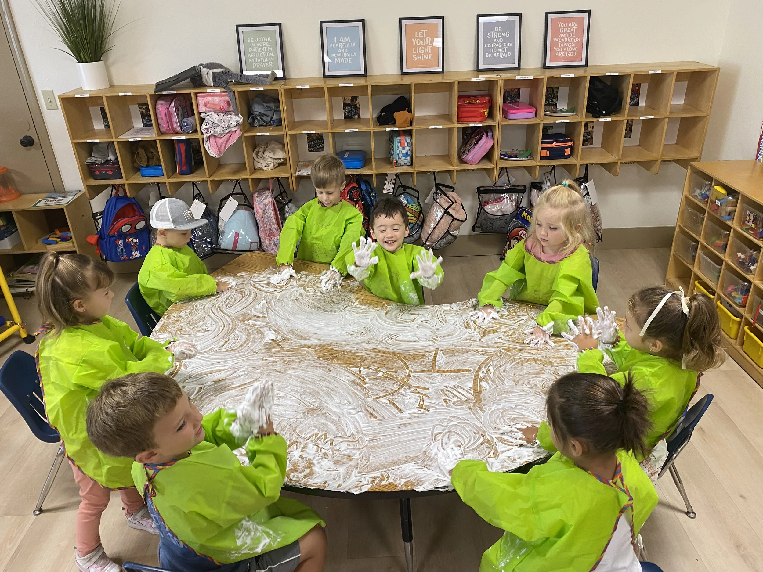 Seven children wearing bright green smocks are sitting around a table covered in soap foam, happily playing with the foam. The classroom has cubbies with backpacks and supplies, and framed motivational quotes on the wall.