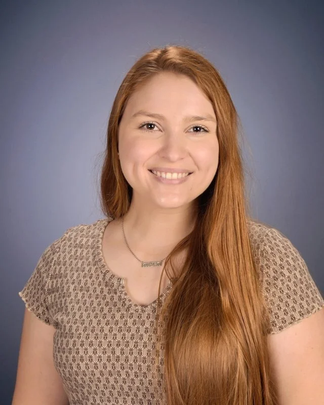 A young woman with long red hair wearing a patterned beige top and a silver necklace, smiling at the camera against a neutral background.
