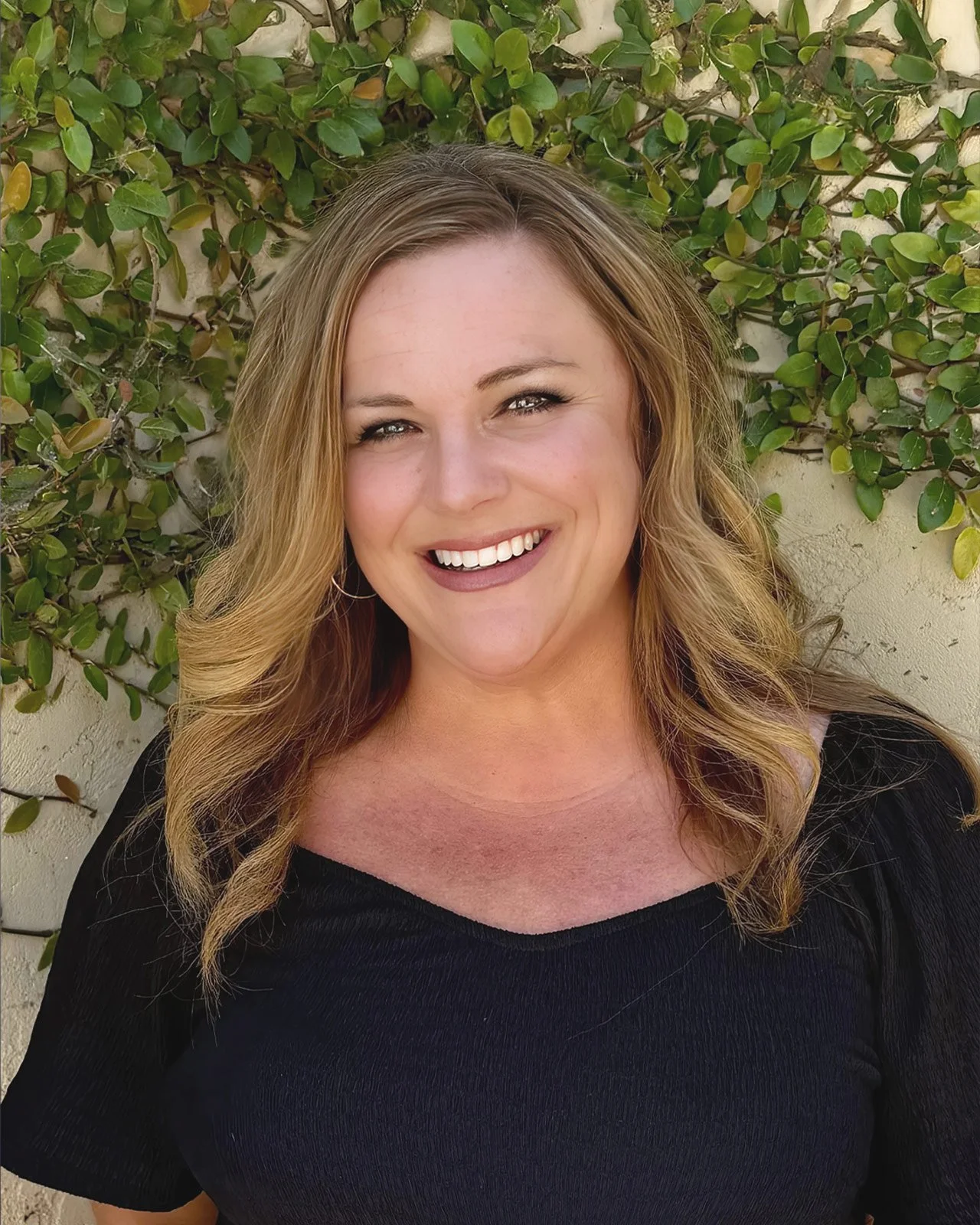 A woman with blonde wavy hair smiling outdoors in front of green leafy plants.
