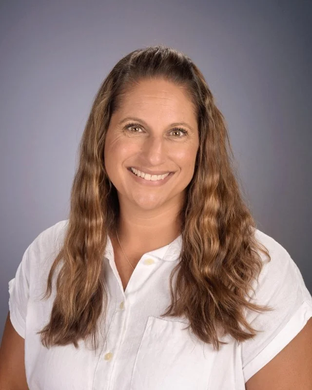 Smiling woman with long wavy brown hair wearing a white shirt against a neutral gray background.