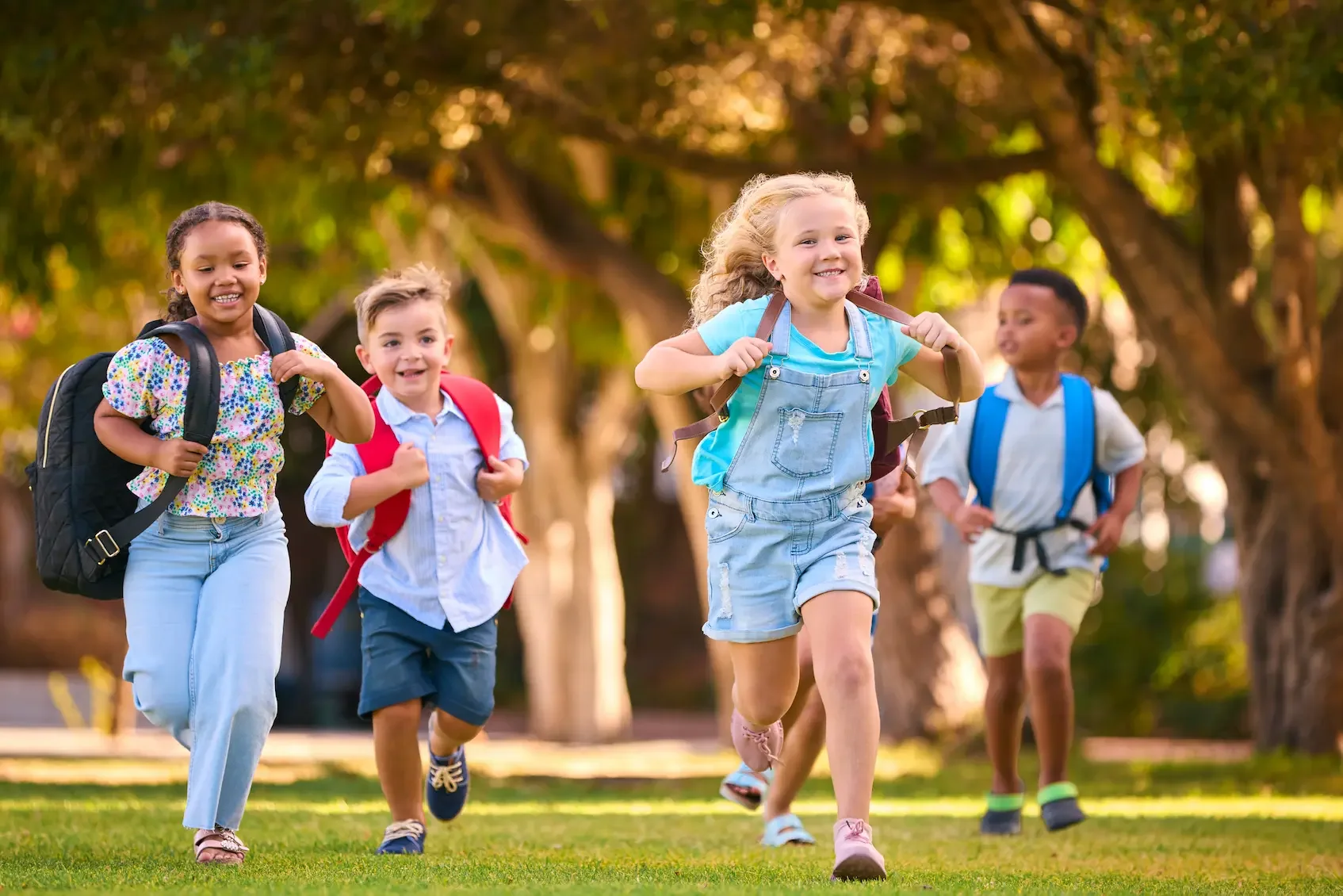 Four children with backpacks running on a grassy area in a park with trees in the background, smiling and enjoying themselves.