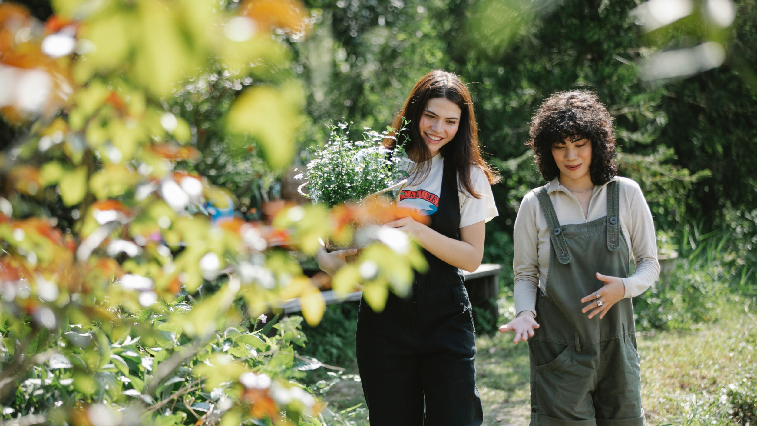 Two women walking through a lush garden, one holding a potted flowering plant, smiling and engaging in conversation.