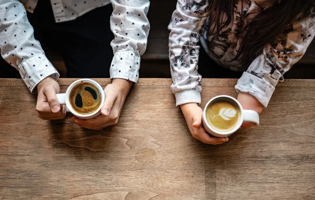 Two children sitting at a wooden table, each holding a coffee mug. One mug contains black coffee, and the other has latte art.